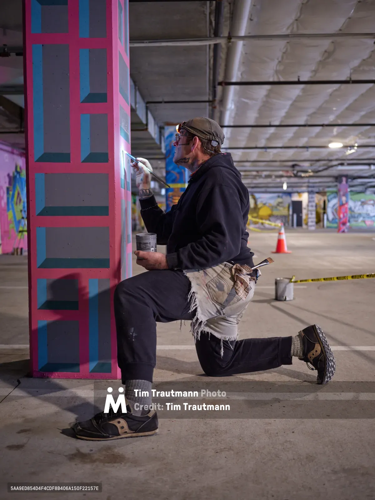 Portland artist and bike activist Rabbitt kneels in focused concentration as he meticulously applies paint to a vibrant geometric mural on a concrete pillar within the Clay Creative building's parking garage. His signature style emerges in bold pink and blue geometric patterns against the industrial backdrop of exposed ceiling beams and concrete surfaces. The intimate moment captures the transformation of urban infrastructure into artistic expression, with paint cans and brushes scattered nearby as tools of creative rebellion.