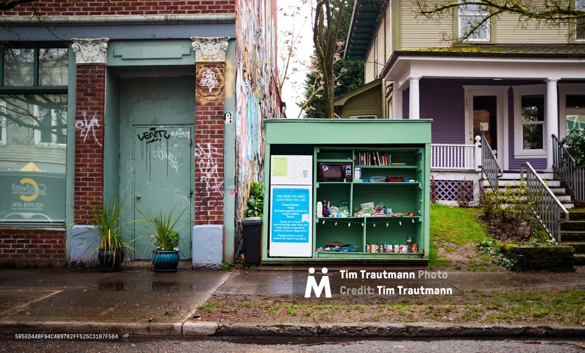 A vibrant lime-green community refrigerator stands as a beacon of mutual aid on Northeast Alberta Street in Portland's Vernon neighborhood. The weathered sidewalk scene captures the intersection of grassroots activism and urban community care, where the colorful fridge contrasts against graffitied brick walls and eclectic residential architecture. Overcast Pacific Northwest light filters through bare winter branches, illuminating this tangible expression of neighborhood solidarity and food justice.