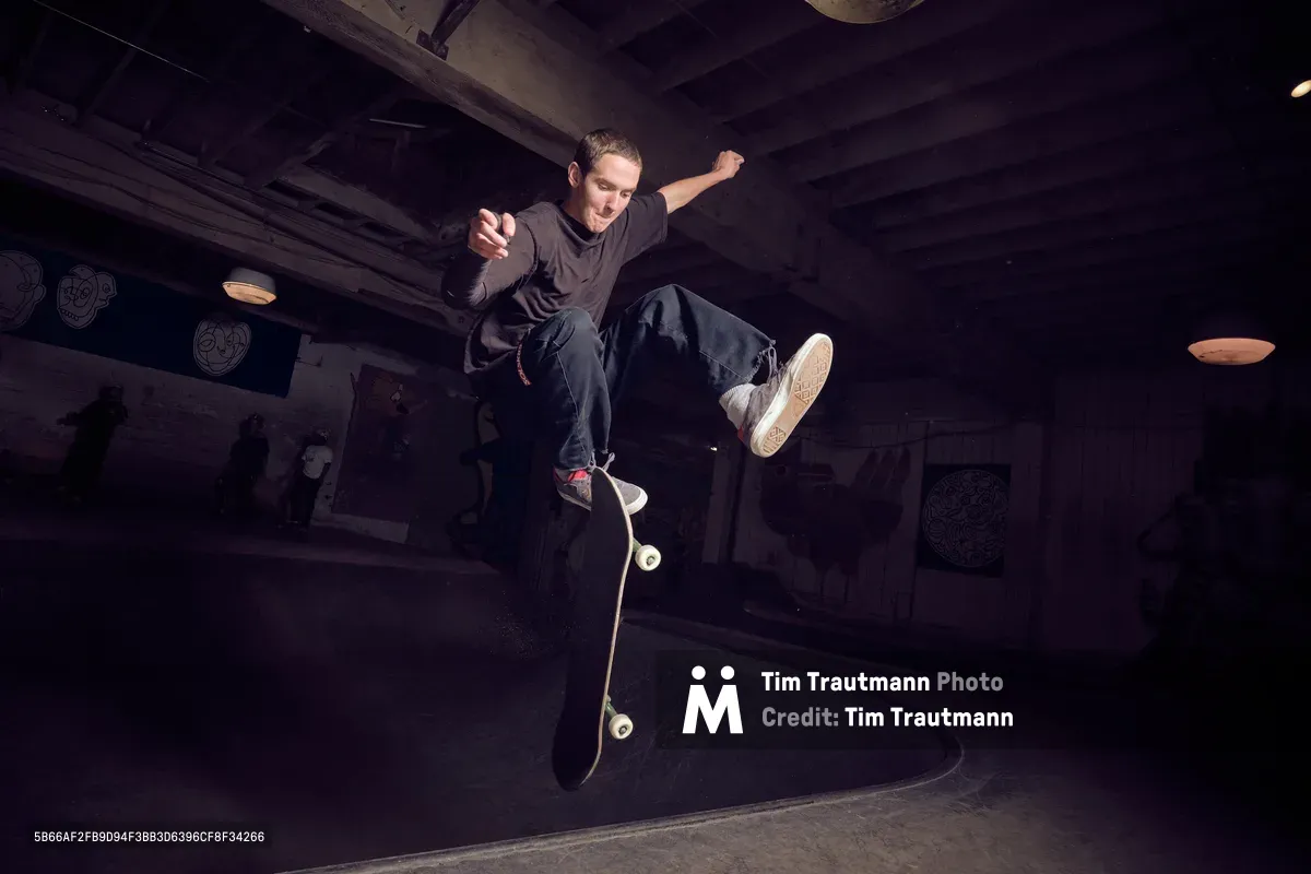 A young skateboarder launches into a dynamic kickflip above the concrete bowl at Commonwealth Skateboarding in Portland's Buckman neighborhood. The dramatic low-angle perspective captures him suspended mid-trick against the shadowy industrial ceiling, his dark clothing silhouetted by moody artificial lighting. The weathered concrete transitions and exposed beams create an authentic underground atmosphere that defines Portland's skateboarding culture.