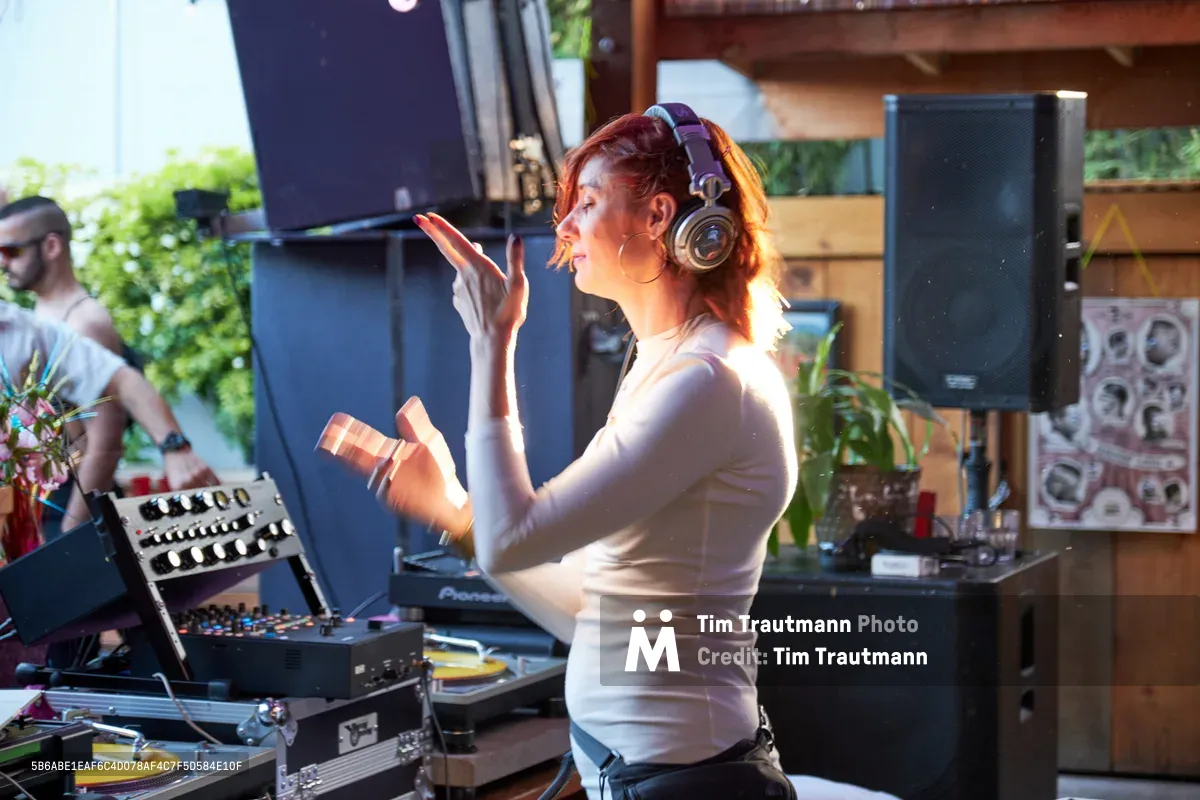 DJ Doctor Dakar commands the decks at White Owl Social Club during the Your Sunday Best season opener, her arms raised in animated gesture as she curates the afternoon's sonic journey. Golden sunlight streams through the venue's windows, illuminating her focused expression beneath professional headphones while vinyl records and synthesizer equipment create an intimate electronic music sanctuary. The Portland day party atmosphere buzzes with creative energy as patrons gather in the background among lush greenery and industrial-chic architecture.