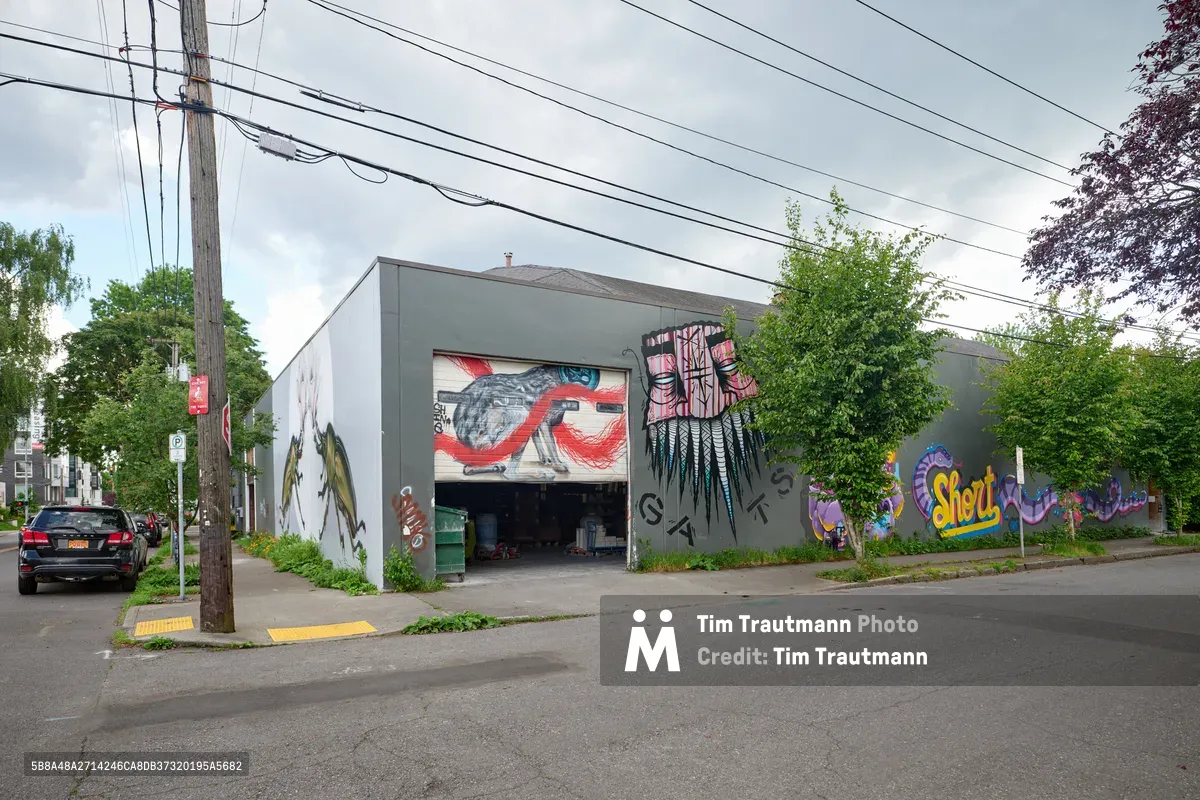 A weathered industrial warehouse in Portland's Kerns neighborhood serves as an expansive canvas for street art, its gray concrete facade adorned with vibrant murals including Native American-inspired imagery and contemporary graffiti tags. Power lines stretch across an overcast sky while mature trees frame the utilitarian structure, creating a quintessential Pacific Northwest urban landscape where industrial heritage meets artistic expression.