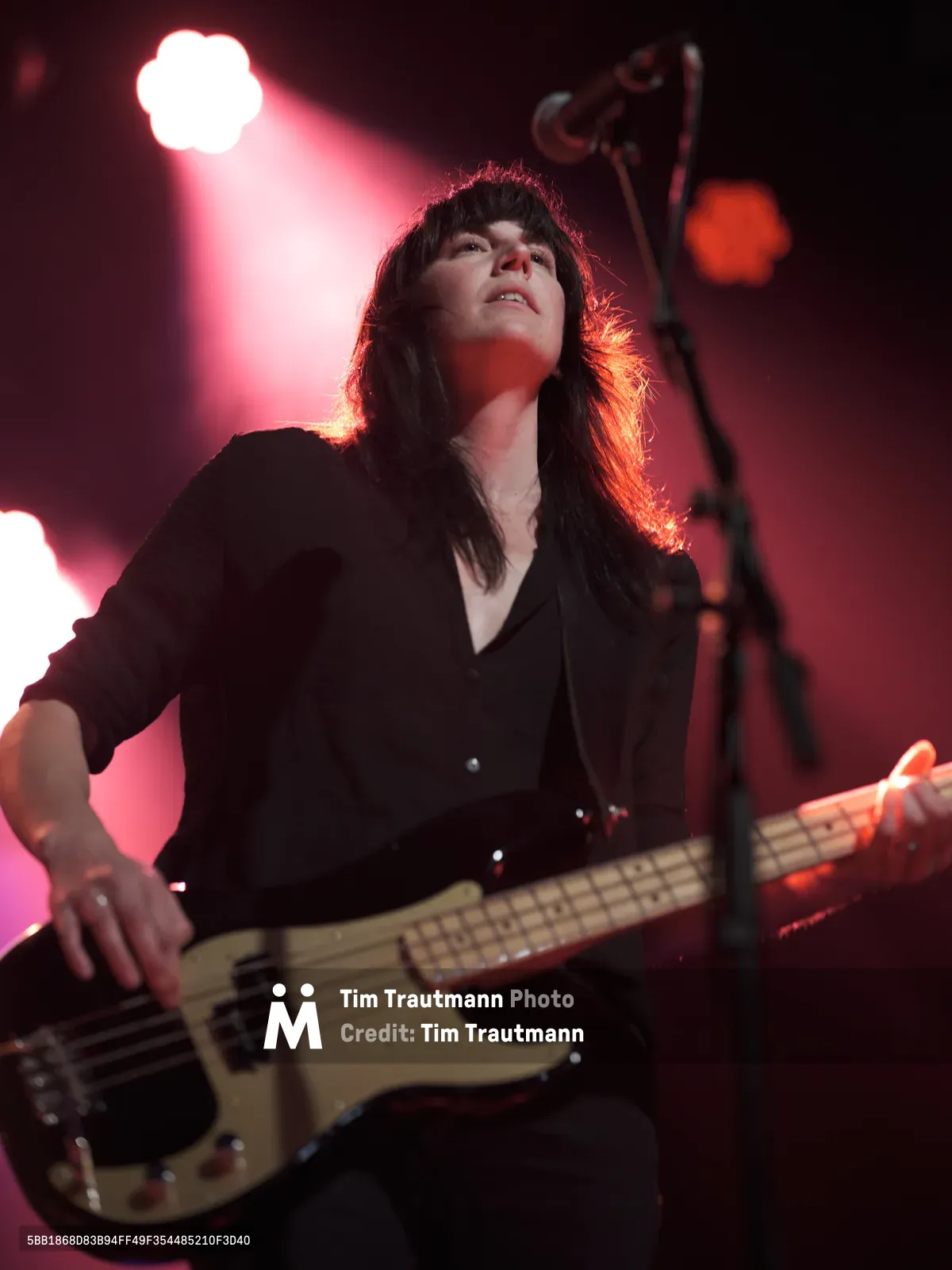 Emma Richardson of Band of Skulls performs with intense concentration at Webster Hall during the band's 10 Year Anniversary Tour. Crimson and amber stage lights create a dramatic silhouette around her flowing dark hair as she grips her cream-colored bass guitar. The low-angle perspective captures her powerful stage presence against the moody lighting of Manhattan's iconic music venue.