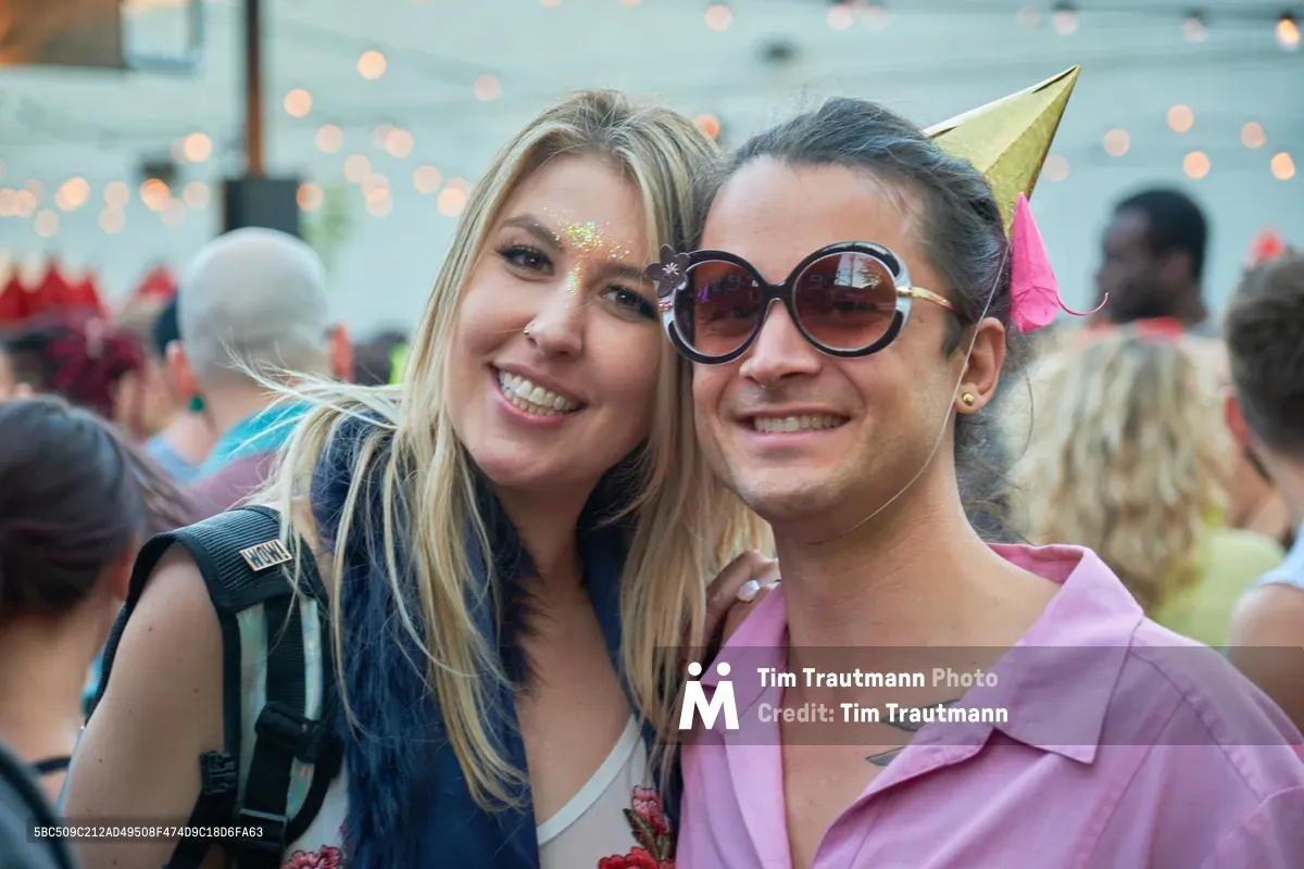 Two young women embrace with radiant smiles at Your Sunday Best day party in Portland's White Owl Social Club. The blonde woman on the left sparkles with golden glitter adorning her face, while her brunette companion sports oversized round sunglasses and a whimsical party hat with pink tassel. Behind them, the warm bokeh of string lights creates an intimate festival atmosphere as other revelers dance in the softly blurred background.