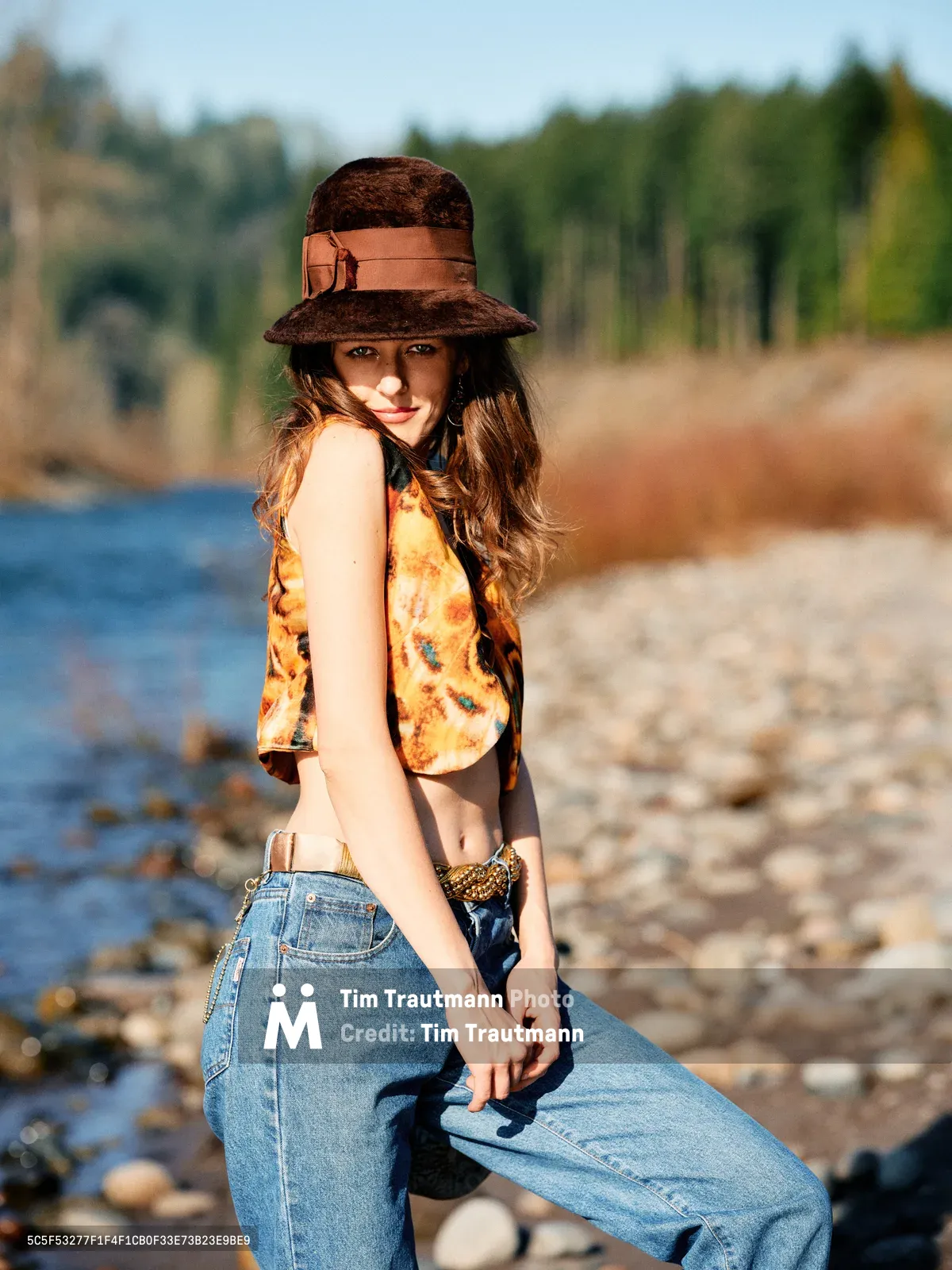 A young woman with flowing brunette hair poses confidently along the rocky riverbed of an Oregon river, wearing a distinctive brown felt hat with leather band, vintage-inspired floral crop top, and high-waisted denim. The golden hour light creates warm shadows across her face while the Columbia River Gorge's forested hillsides provide a dreamy backdrop. Her relaxed stance and gentle smile capture a moment of carefree summer exploration along the river bed.