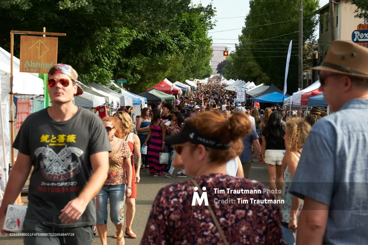 Hundreds of festival-goers meander through a bustling street market along North Mississippi Avenue in Portland's Boise neighborhood. White vendor tents line both sides of the closed street, creating a vibrant corridor filled with browsing shoppers under the dappled shade of mature trees. The late afternoon sunlight filters through leafy canopies, illuminating the diverse crowd as they explore local artisan booths and food vendors in this quintessential Pacific Northwest community gathering.