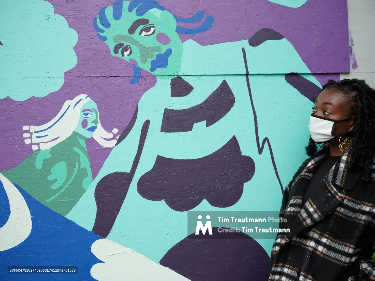 Artist Naomi Likayi stands beside her vibrant mural painted on the boarded Portland World Trade Center, wearing a white face mask and patterned coat. The artwork features flowing figures rendered in soft blues, purples, and mint greens against a rich purple background, creating a dreamlike narrative across the temporary urban canvas. Her contemplative gaze and proximity to the work establishes an intimate connection between creator and creation. The street art transforms the utilitarian boarding into a window of imagination and color.