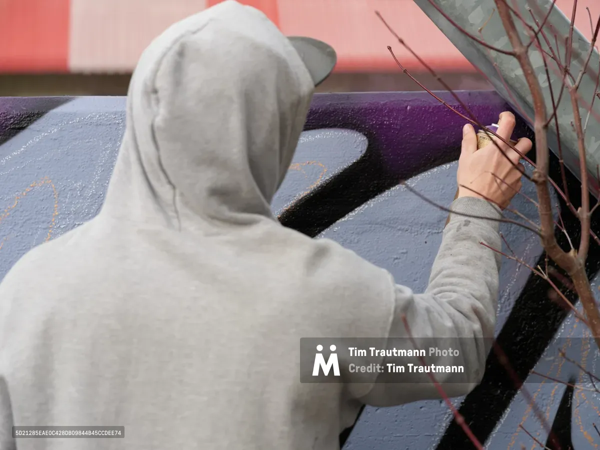 Travis Fields, known as Campographic, works intently on a vibrant mural at the Clay Creative Building in southeast Portland's Hosford-Abernethy neighborhood. Wearing a gray hoodie, the artist reaches toward purple and black paint strokes while delicate bare branches frame the foreground. The soft afternoon light illuminates the textured wall surface, capturing the meditative focus of artistic creation in this converted industrial arts space.