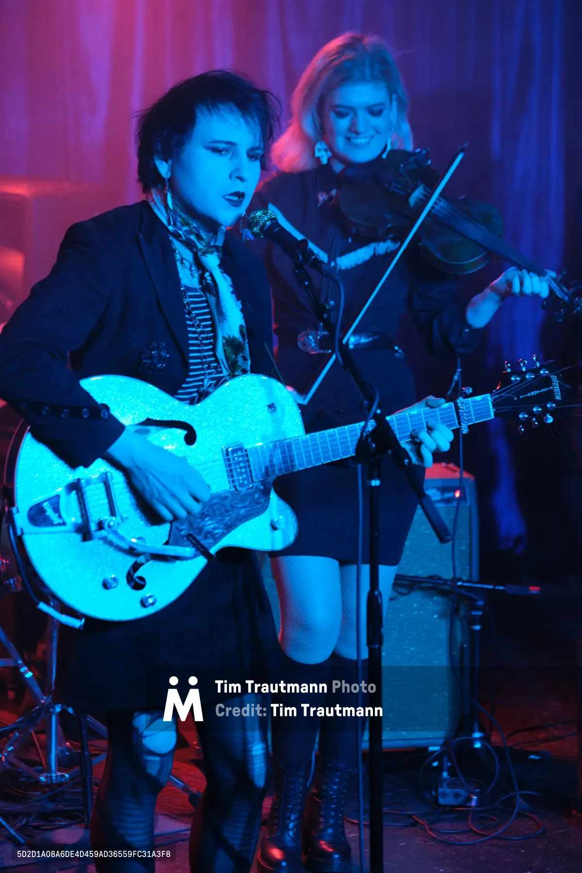 Charlotte McCaslin grips her white hollow-body guitar with fierce concentration while Faith Grossnicklaus draws her violin bow behind her, both performers bathed in the saturated purple and blue stage lights of Portland's legendary Dante's venue. The intimate underground atmosphere crackles with electric energy as the duo delivers their signature blend of rock and classical instrumentation. McCaslin's dark tousled hair and vintage-inspired outfit contrast sharply with the ethereal glow of the stage lighting, while Grossnicklaus remains partially obscured in the moody shadows.