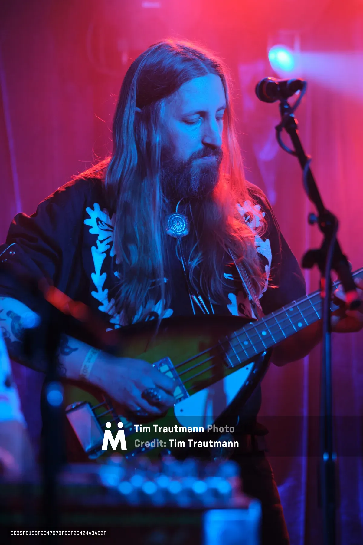 Tobias Berblinger of Roselit Bone emerges from a dreamscape of magenta and cyan stage lighting, his flowing blonde hair and full beard catching the electric glow as he cradles his bass guitar. The intimate venue pulses with atmospheric haze, transforming the musician into a spectral figure lost in concentration. His dark shirt adorned with white botanical prints creates striking contrast against the saturated colors washing over Dante's legendary stage in Portland's Old Town.