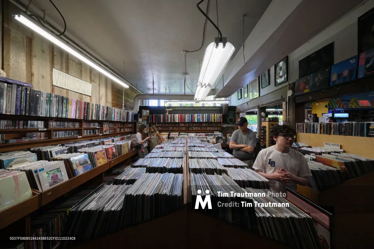 Inside Music Millennium on East Burnside Street in Portland, Oregon, customers browse through extensive collections of vinyl records organized in wooden bins that stretch down the center aisle. The independent record store's industrial ceiling features exposed fluorescent lighting, while walls lined with albums and CDs create a maze of musical discovery. Three patrons move through the space, each absorbed in their search through thousands of records, embodying the tactile ritual of vinyl hunting in this legendary Portland music institution.