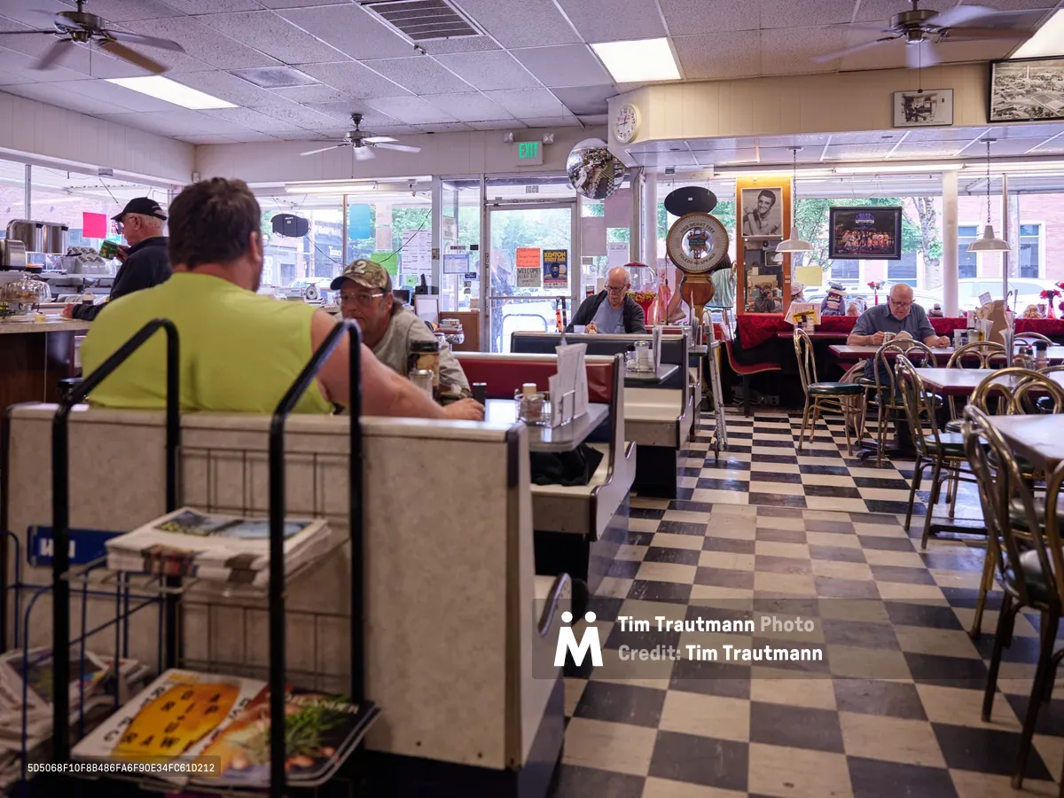 The weathered checkerboard linoleum tells stories beneath the gentle hum of ceiling fans at Pattie's Home Plate Café in Portland's Saint Johns neighborhood. Regulars occupy their familiar stations—a man in a lime safety vest studies his newspaper at the counter while others settle into red vinyl booths bathed in soft fluorescent light. The diner's lived-in atmosphere speaks to decades of community gathering, where morning coffee and conversation flow as predictably as the Oregon rain outside the storefront windows.