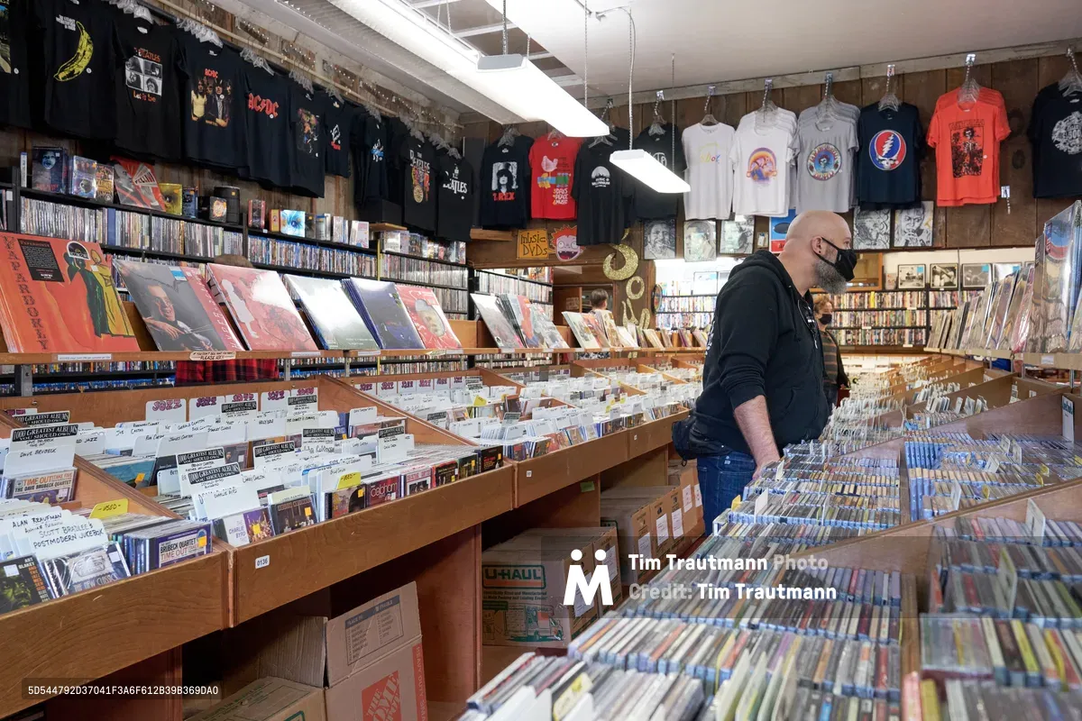 A bearded man in a black hoodie and face mask browses through wooden crates overflowing with vinyl records at Music Millennium on Burnside Street during Record Store Day 2022. The warm fluorescent lighting illuminates the cramped aisles lined with thousands of albums, creating an intimate atmosphere of musical discovery. Band t-shirts hang overhead like colorful flags, while shelves stretch toward exposed ceiling beams packed with decades of recorded music history.