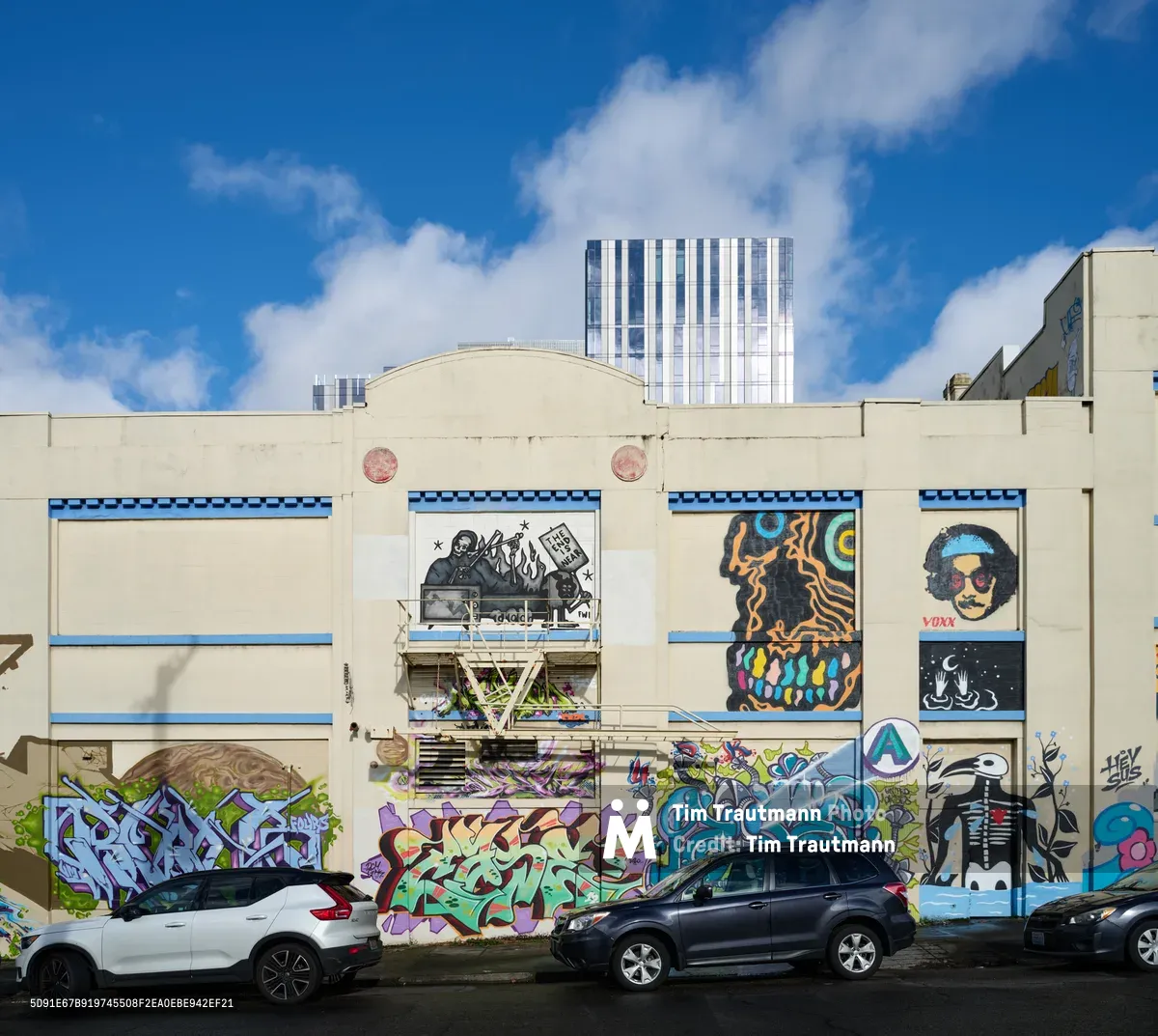 A vibrant tapestry of commissioned murals adorns the cream-colored facade of a former Salvation Army warehouse on Southeast Ash Street in Portland's Central Eastside district. The eclectic street art collection features bold graffiti-style pieces, character portraits, and abstract designs framed by distinctive blue trim, creating a dynamic gallery against the industrial backdrop. Modern SUVs parked along the street provide scale to this transformed urban landscape, while gleaming office towers pierce the partly cloudy sky beyond.