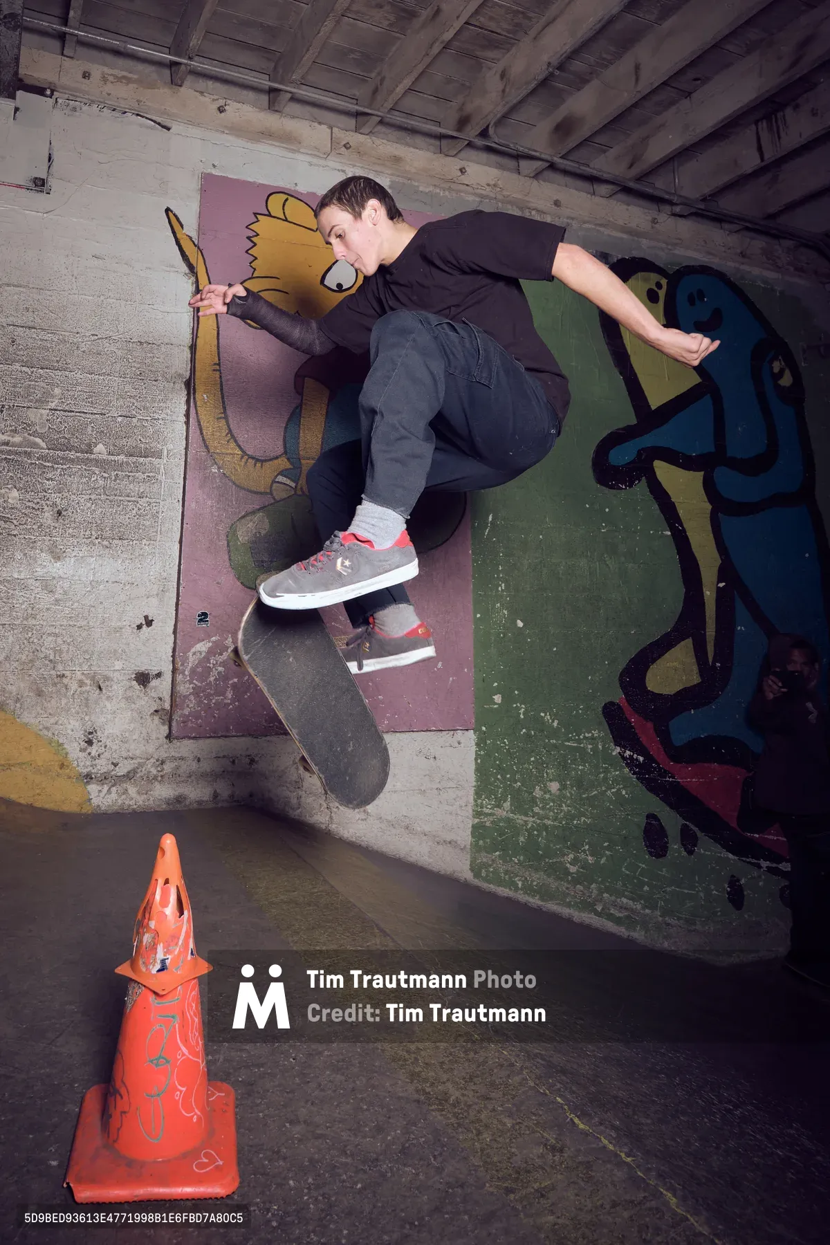 A skateboarder captures air against a vibrant graffiti-covered wall in the underground confines of Commonwealth Skateboarding in Portland's Buckman neighborhood. The raw concrete space, punctuated by exposed ceiling beams and industrial elements, creates a gritty urban canvas where street art meets athletic expression. An orange traffic cone sits sentinel on the worn floor, while the skater's form is frozen mid-trick against the colorful backdrop of cartoon-like murals.