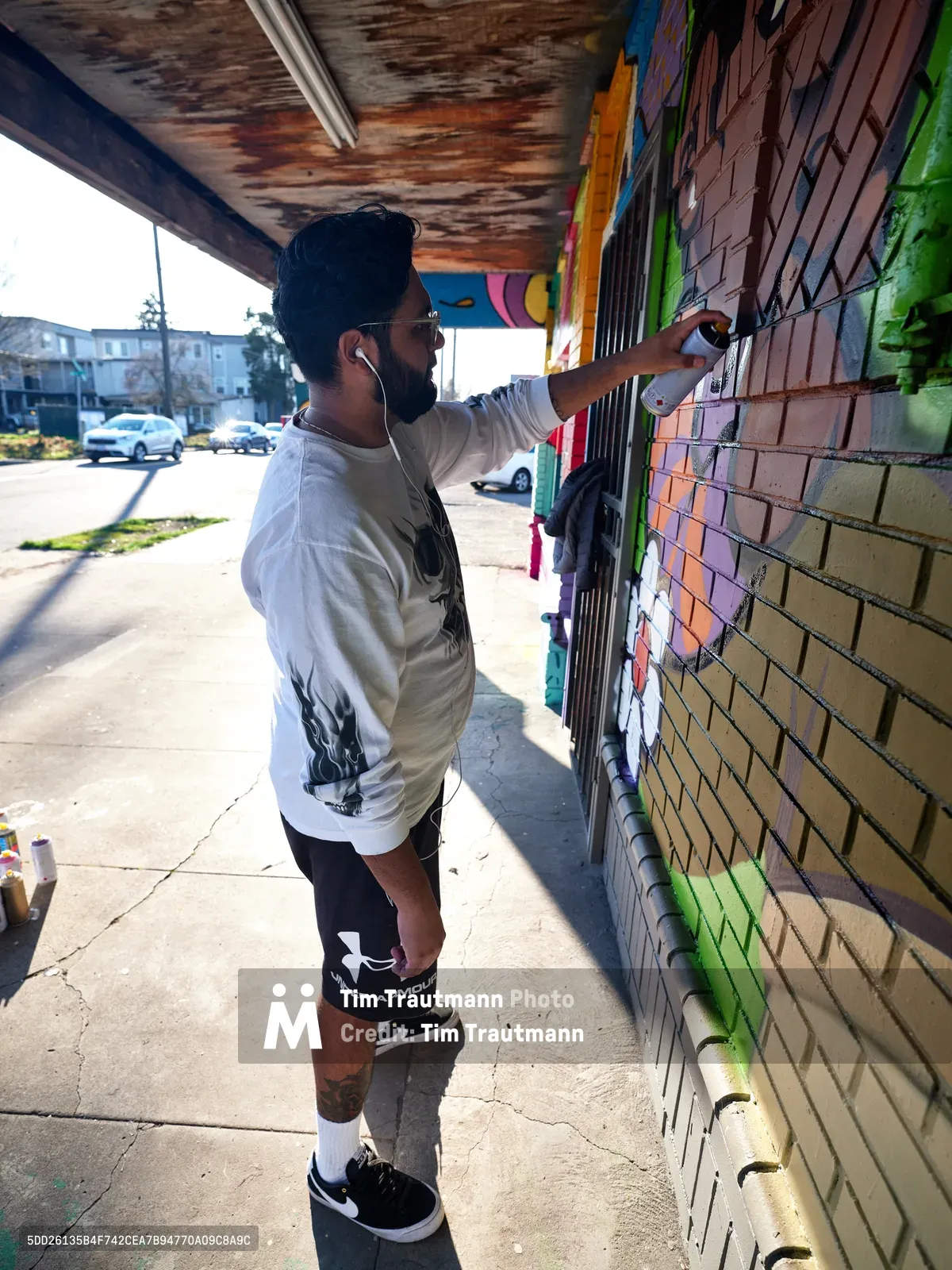 Muralist Heysus concentrates intensely as he adds precise details to a vibrant community mural on a brick wall at the intersection of Killingsworth and Northeast 15th Avenue in Portland's Alberta district. The weathered overhang casts dramatic shadows across the concrete sidewalk while afternoon light illuminates the artist's focused expression and the explosion of colors transforming the urban surface. His white long-sleeve shirt contrasts against the rainbow spectrum of spray paint emerging from his careful hand movements.