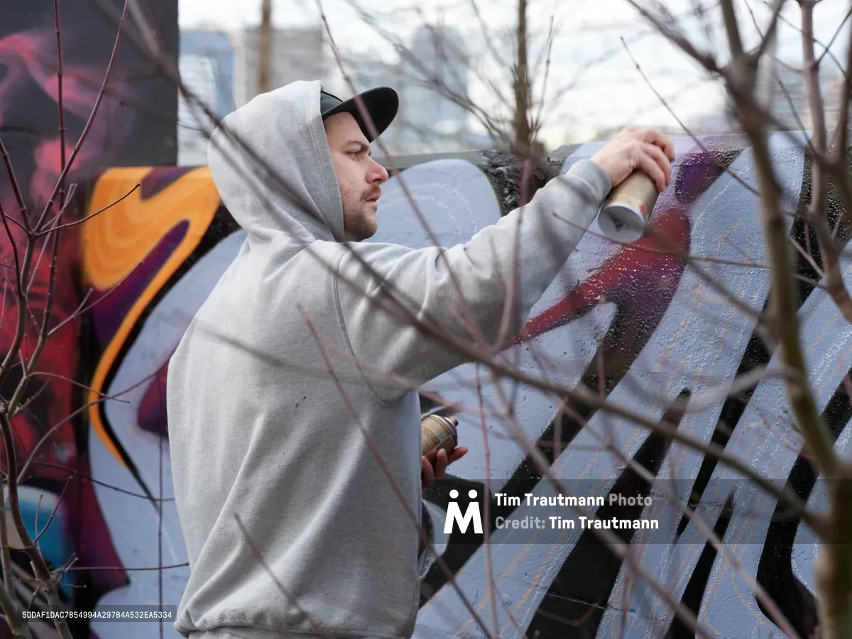 Travis Fields, known as Campographic, concentrates intently as he applies purple spray paint to a concrete wall at the Clay Creative Building in southeast Portland. The hooded artist works methodically through bare winter branches that create natural framing in the foreground, while colorful existing graffiti provides a vibrant backdrop. Soft overcast light illuminates this intimate moment of urban artistry in the Hosford-Abernethy neighborhood.