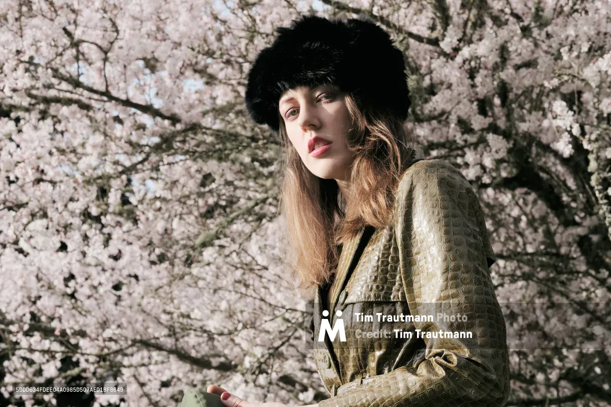 Model Rhianna Atwood gazes thoughtfully beneath a canopy of flowering cherry trees at Mount Tabor in Portland, Oregon. The golden hour light filters through the abundant white blossoms, creating a dreamy hanami atmosphere. She wears a luxurious olive-toned crocodile textured coat paired with a striking black fur hat, her flowing blonde hair catching the warm spring light. The composition captures the ephemeral beauty of sakura season against Portland's urban parkland backdrop.