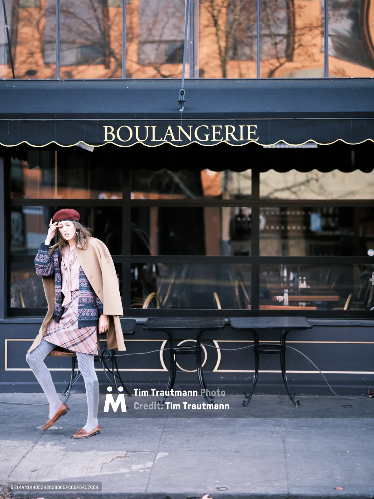 A young woman in layered autumn attire pauses mid-stride along Northeast Alberta Street in Portland, Oregon, her burgundy beret and mixed-pattern ensemble creating a study in effortless urban sophistication. The scene unfolds before a classic French boulangerie with its traditional black awning and golden lettering, while wrought iron café tables sit empty on the sidewalk. Golden hour light bathes the storefront's large windows, casting warm reflections that complement the subject's camel coat and plaid scarf, capturing the essence of Portland's vibrant Alberta Arts District.