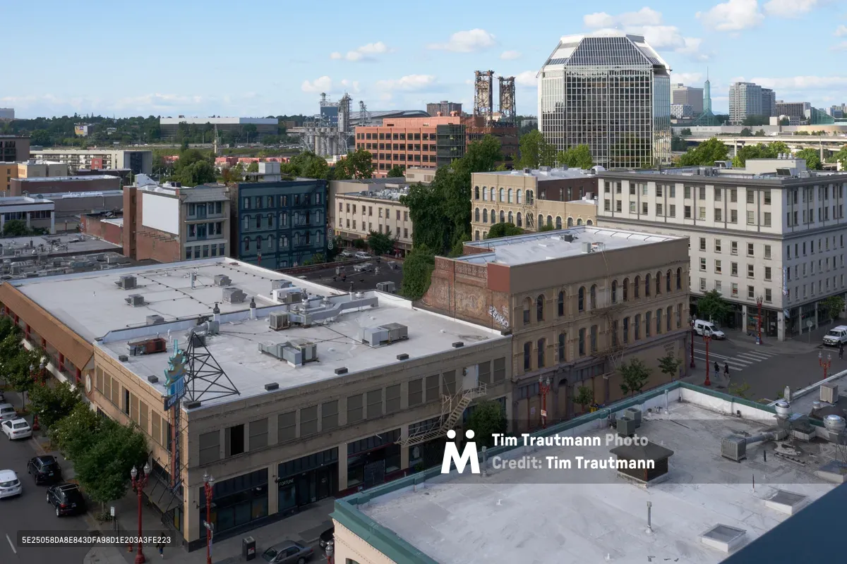 An elevated perspective captures the historic architecture of Portland's Chinatown district on a partly cloudy afternoon. The foreground showcases weathered brick buildings with flat rooftops dotted with HVAC equipment, while mid-century structures with distinctive facades stretch toward the horizon. In the distance, gleaming downtown high-rises pierce a soft blue sky scattered with white cumulus clouds, creating a compelling juxtaposition between the neighborhood's intimate scale and the city's vertical ambitions.