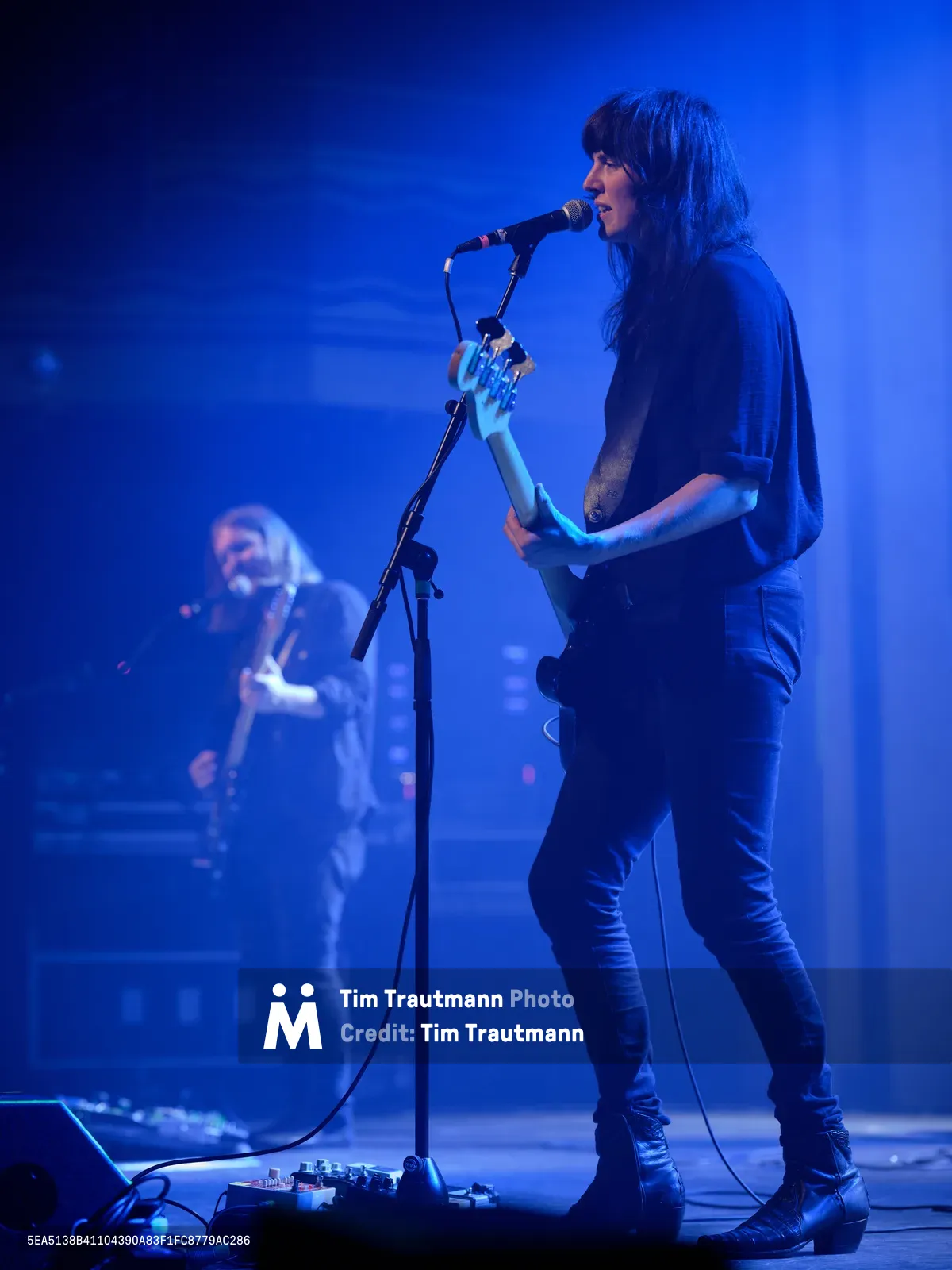 Emma Richardson of Band of Skulls commands the stage at Webster Hall during the band's 10 Year Anniversary Tour, her silhouette illuminated against a deep cobalt backdrop. The intimate venue's atmospheric lighting casts dramatic shadows as she performs alongside bandmate Russell Marsden, who remains partially visible in the misty background. Richardson's focused expression and dynamic posture capture the raw energy of live indie rock performance in Manhattan's historic East Village music venue.