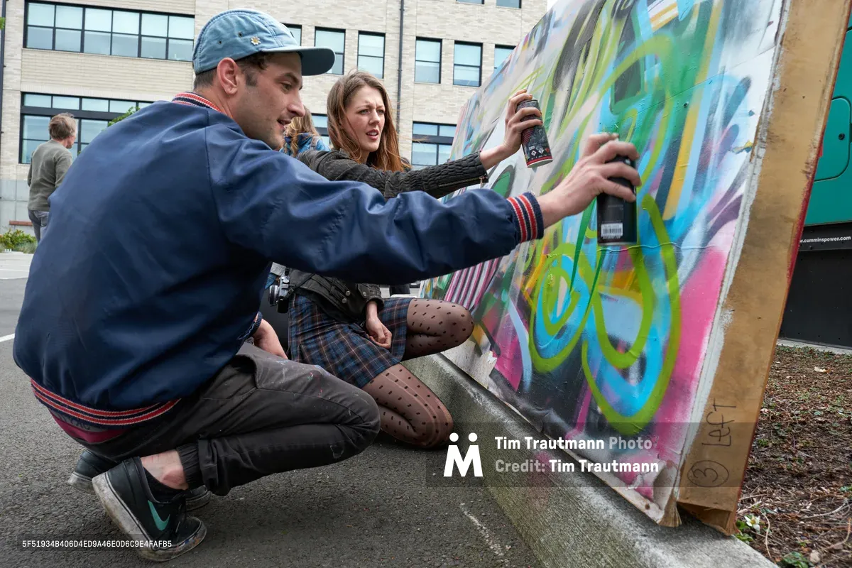 Galen Malcolm of the Portland Street Art Alliance crouches beside a vibrant canvas, spray can in hand, while demonstrating live graffiti techniques to an engaged woman in polka-dotted tights. The scene unfolds against the backdrop of Portland's Taylor Electric Blocks, where colorful street art education meets community engagement. Warm afternoon light illuminates the collaborative moment as the instructor guides his student through the rhythmic motions of aerosol artistry.