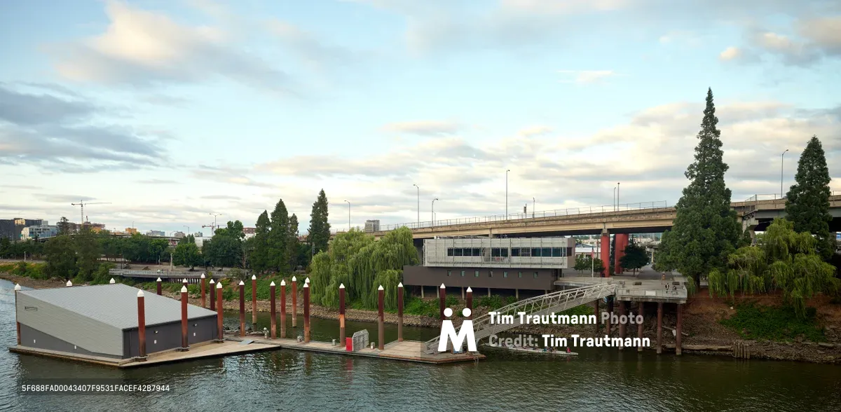 Portland Fire & Rescue Station 21 sits anchored in the Willamette River beneath the Southeast Madison Street Bridge, its modern floating structure serving the Central Eastside industrial district. The utilitarian gray building extends over dark waters on wooden pilings, connected to shore by a pedestrian gangway that spans the vegetated riverbank. Towering conifers and weeping willows frame the scene under a brooding Pacific Northwest sky, while the concrete bridge arches overhead, linking the city's urban fabric.