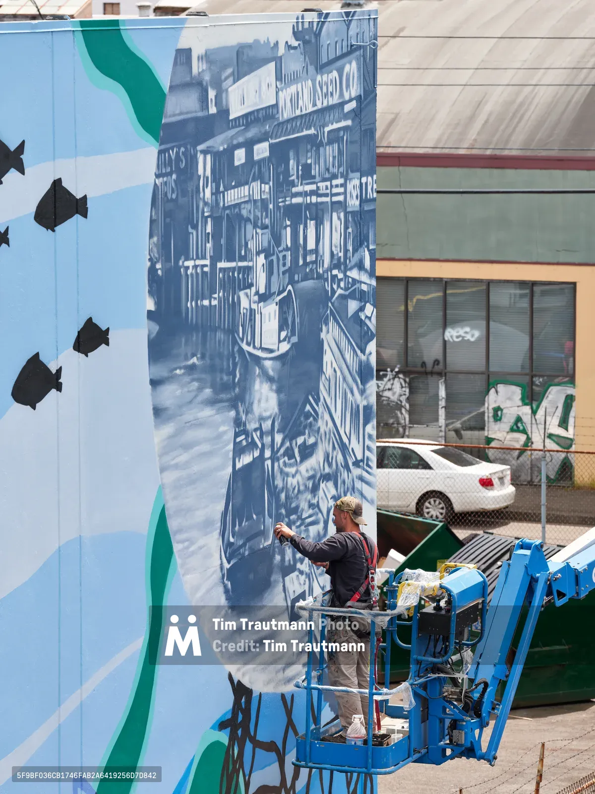Jeremy Nichols and Devin Finley work from a blue scissor lift to paint an elaborate monochromatic riverboat mural on a building wall in Portland's Central Eastside district. The detailed artwork depicts a historic paddle steamer with visible signage including 'Natural Seed Co.', rendered in blue and white tones against the industrial urban backdrop. Black fish silhouettes swim across the adjacent wall section, creating a cohesive aquatic narrative that transforms the concrete surface into a nostalgic river scene.