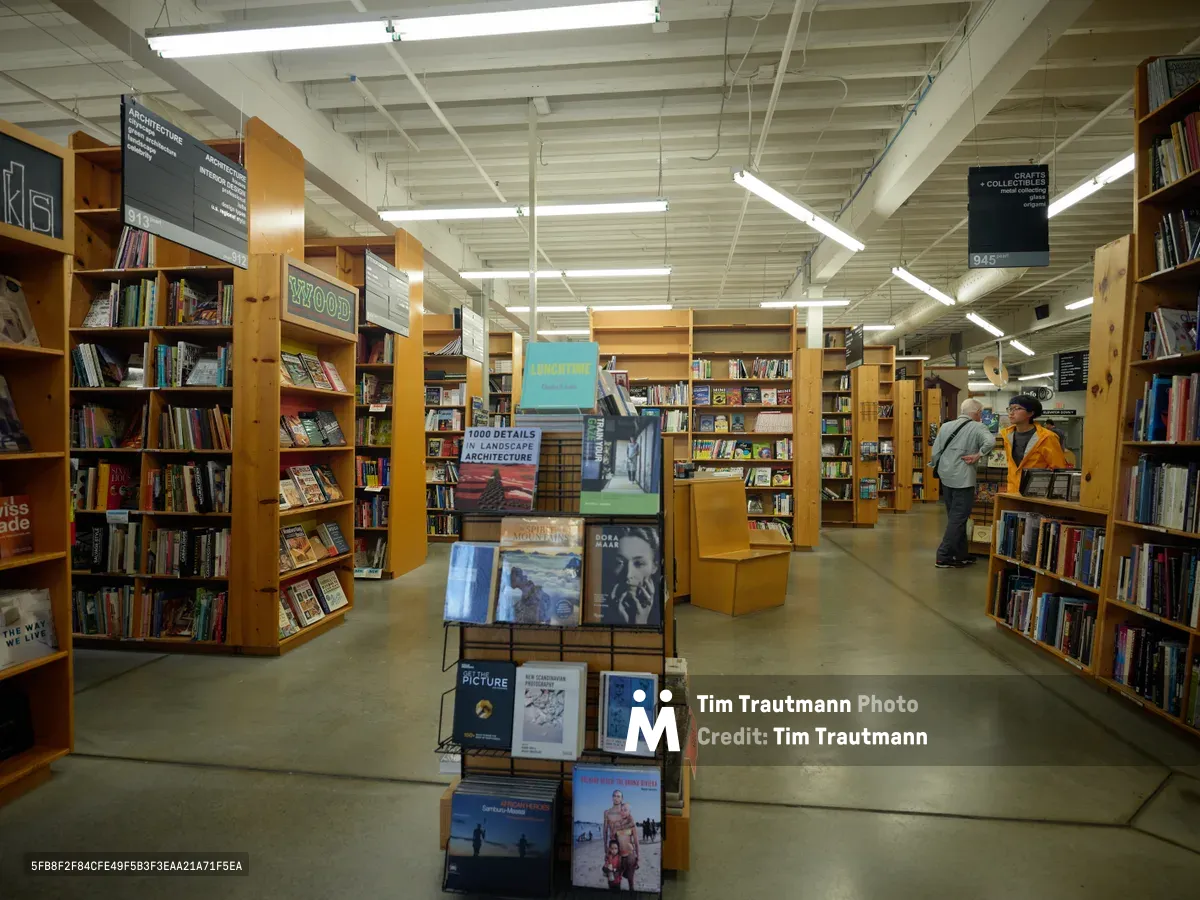 Fluorescent light bathes the towering honey-colored shelves of Powell's City of Books, creating geometric patterns across polished concrete floors. Two visitors in casual attire browse quietly among meticulously organized sections marked by hanging black signage, while a central display showcases architecture and landscape photography books. The exposed ceiling's industrial bones frame this temple of knowledge, where thousands of volumes create a labyrinthine sanctuary for bibliophiles.