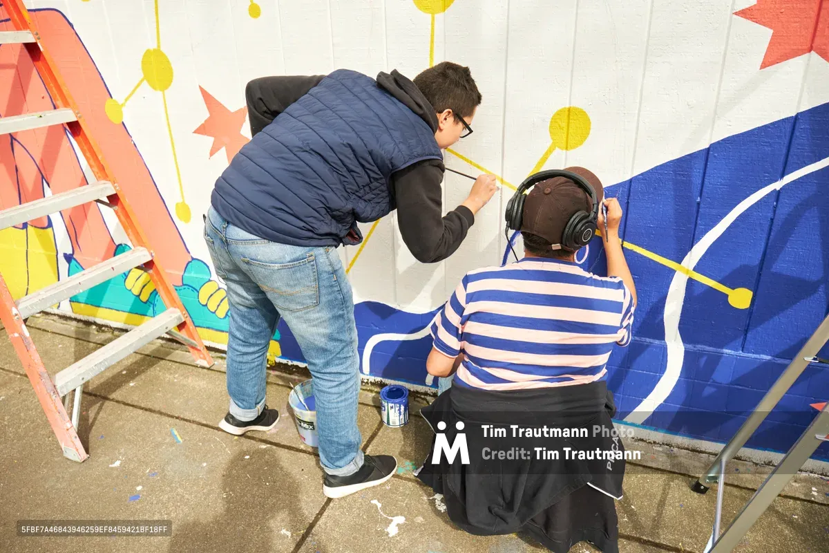 Anke Gladnick and Maria Rodriguez collaborate on a vibrant public mural titled "Let's Talk" at Open Signal in Portland's Eliot neighborhood. The standing artist in navy vest works alongside a seated partner wearing headphones and striped shirt, their brushes bringing bold geometric patterns to life against a white wall. Paint cans and a wooden ladder frame the scene as the duo transforms the community space with radiating circles and angular forms in coral, blue, and golden yellow.