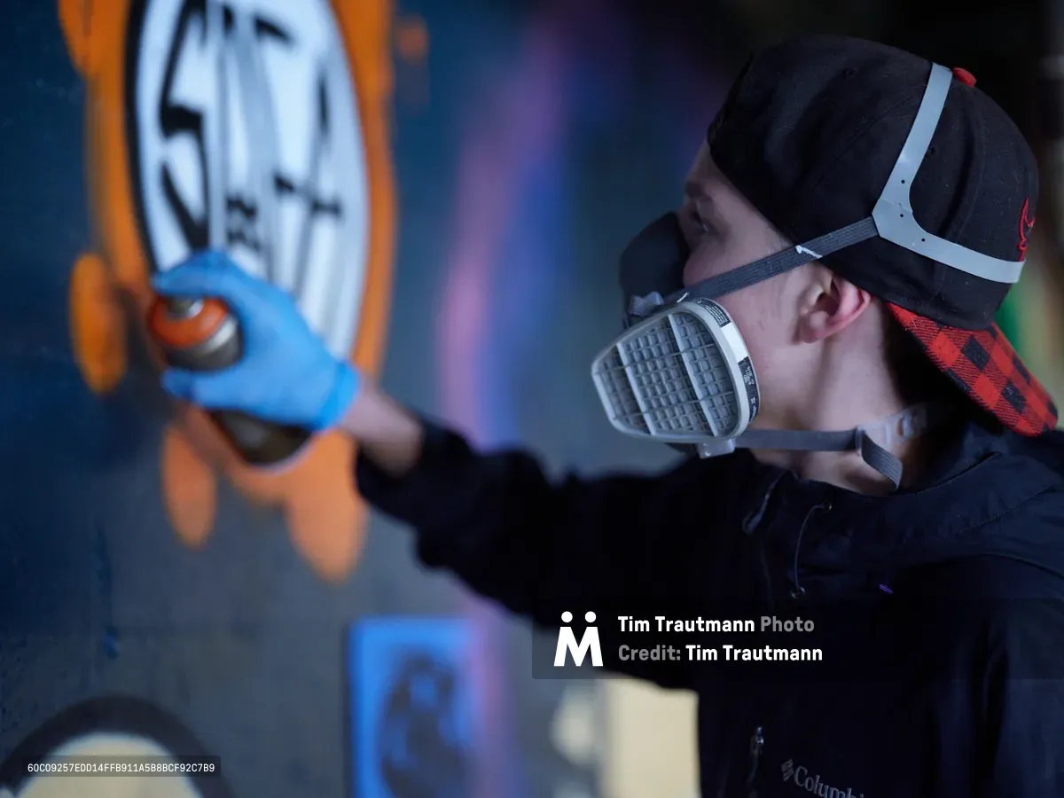 In the atmospheric depths of Clay Creative's parking garage in southeast Portland's Hosford-Abernethy neighborhood, a young artist wearing a protective respirator and backwards cap applies vibrant blue paint to an emerging mural. The scene captures the intersection of urban art and industrial space, with colorful graffiti work illuminated against the concrete walls. Spray paint equipment and artistic materials suggest an active creative process unfolding in this converted creative space.