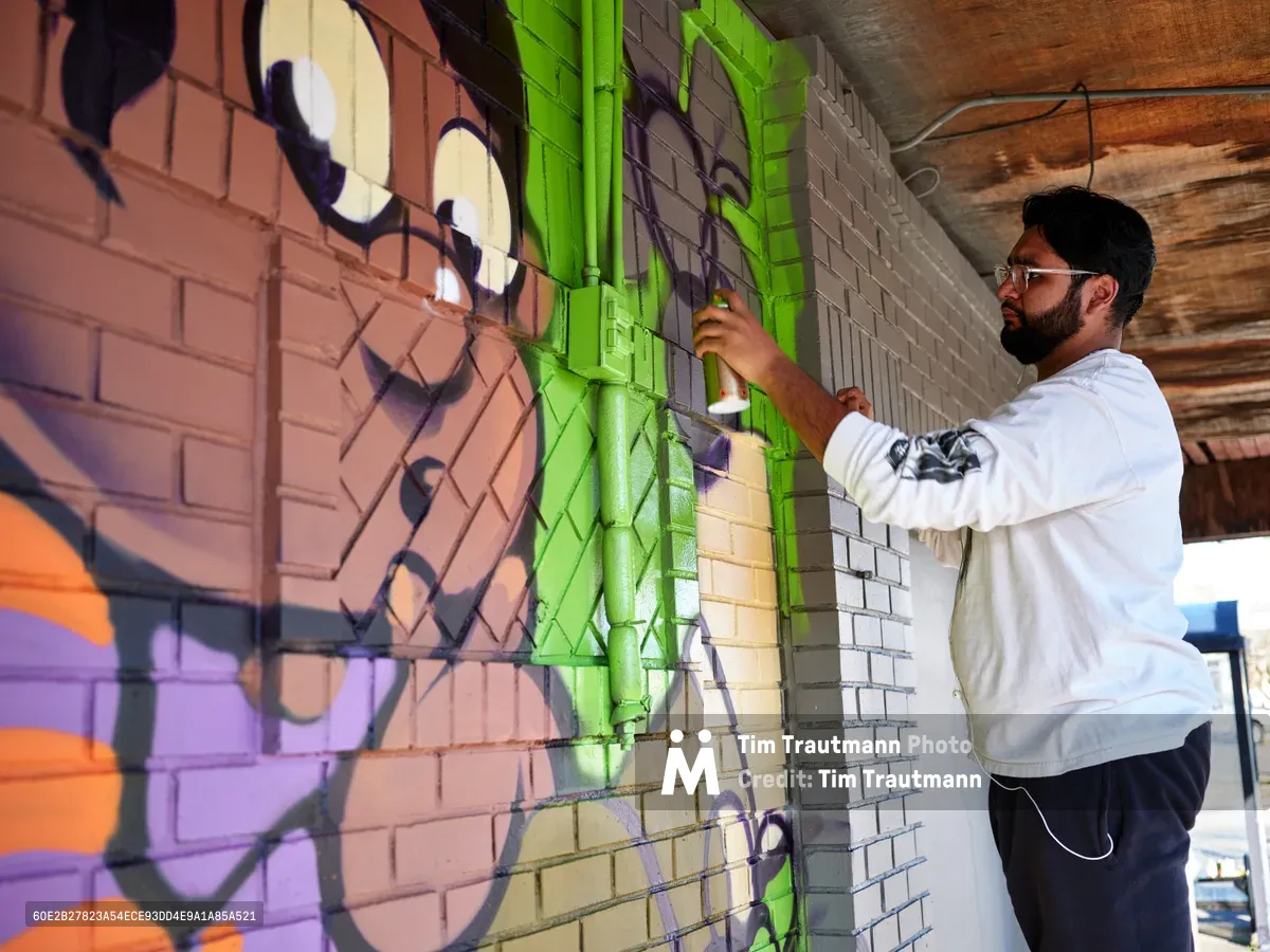 Muralist Heysus concentrates intently as he applies electric lime green paint to a dynamic street art composition on the brick wall of a building in Portland's Alberta neighborhood. The bearded artist, wearing a white hoodie and glasses, works methodically on his colorful creation featuring bold purple, orange, and pink elements that flow across the textured masonry surface. Afternoon light filters through the covered workspace, illuminating both the emerging artwork and the focused determination of the creator.