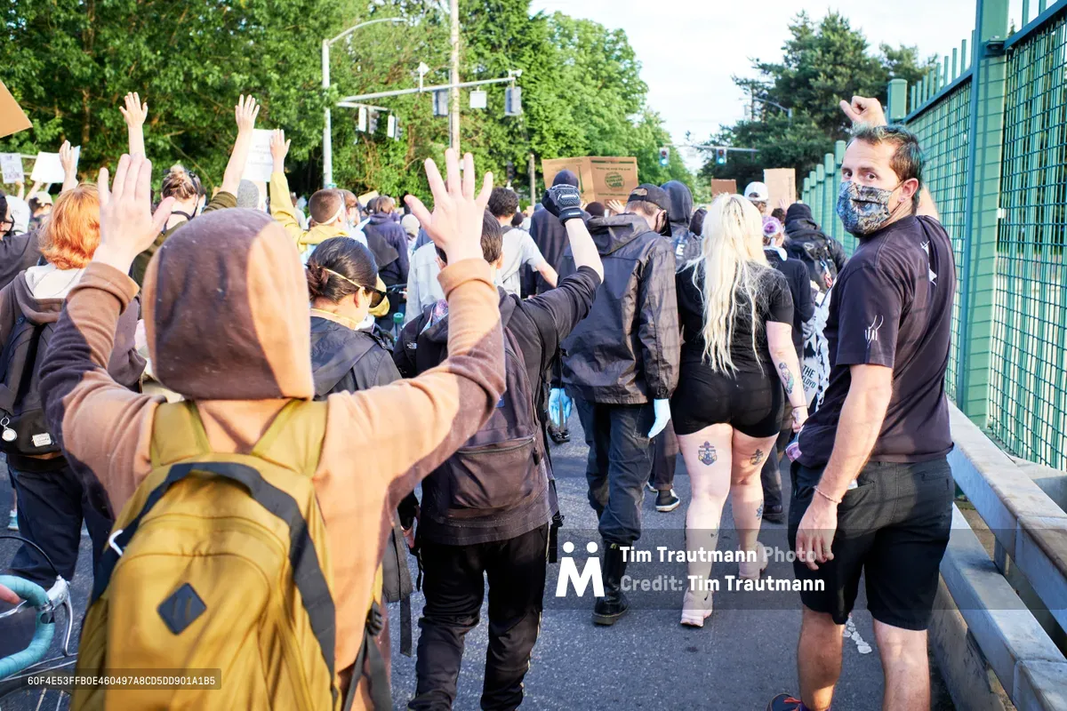 Protesters with raised hands move through Northeast Portland during a Black Lives Matter demonstration following George Floyd's murder. A masked organizer in black clothing stands watchfully beside the green security fence as demonstrators in hooded jackets and backpacks flow past on the asphalt street. The scene captures the solemn determination of the movement, with late afternoon light filtering through the leafy urban canopy above Sullivan's Gulch.