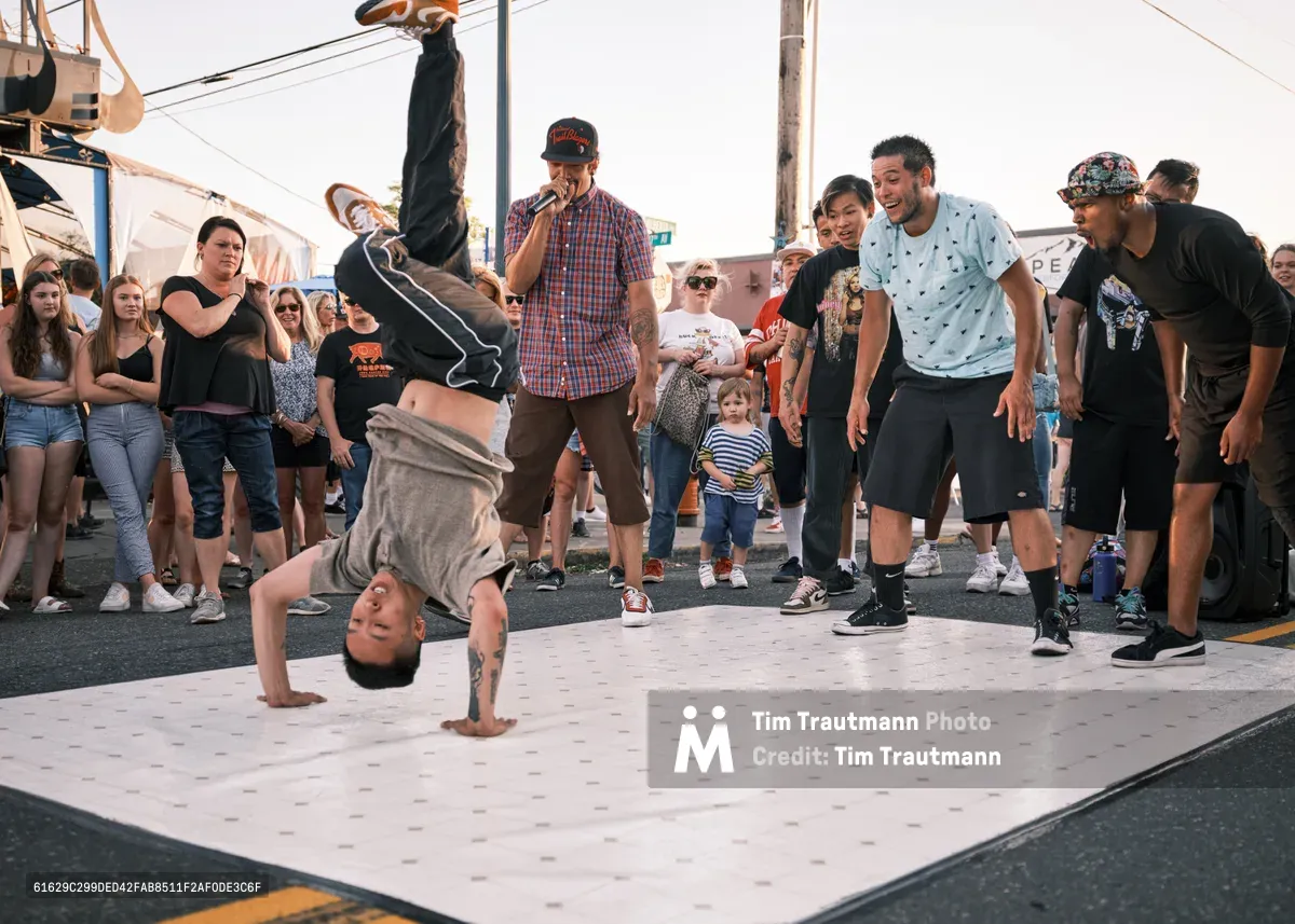 A skilled breakdancer executes a dynamic freeze move on a white mat as an enthusiastic crowd circles around him during Portland's Last Thursday street fair on Alberta Street. The late afternoon light casts warm shadows across the asphalt while spectators of all ages lean in to witness the impromptu performance. Onlookers range from young families to hip-hop enthusiasts, their expressions capturing the magnetic pull of street culture in Northeast Portland's vibrant arts district.
