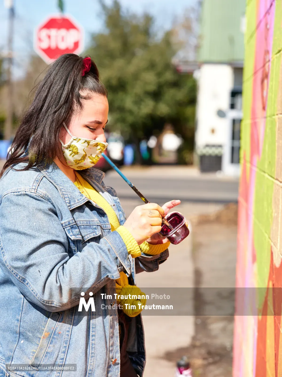 Multidisciplinary artist Vilatose pauses mid-creation while working on 'Jardín Cubana,' a vibrant mural depicting three female figures along Northeast Alberta Street in Portland's renowned Alberta Arts District. Wearing a pineapple-patterned face mask and denim jacket layered over a mustard sweater, she holds painting supplies against the backdrop of the colorful wall. The afternoon light catches the red stop sign and blurred streetscape behind her, capturing a moment of artistic process in one of Portland's most culturally significant neighborhoods.