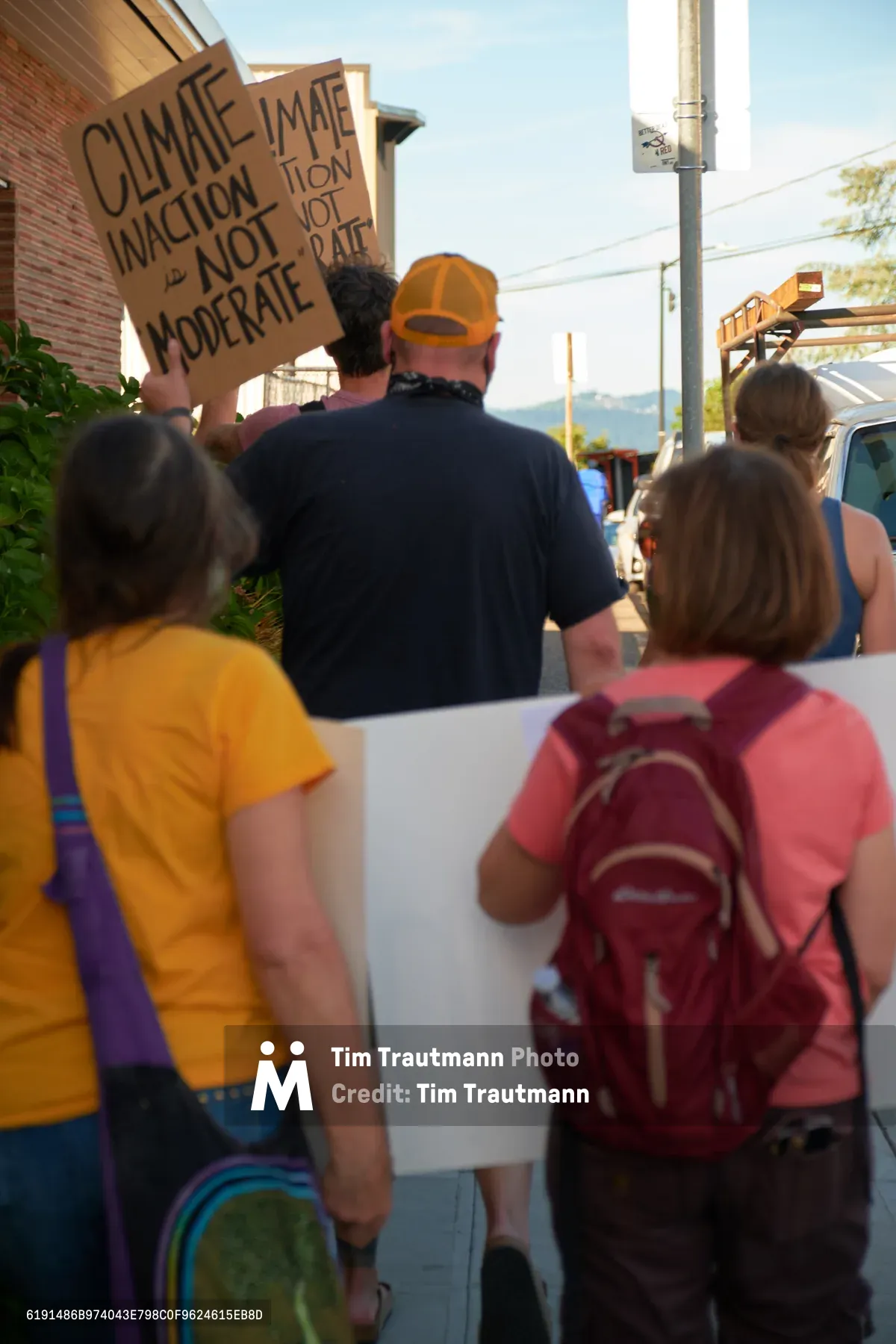 A diverse group of environmental protesters moves through McLoughlin Promenade in Oregon City, their handmade cardboard signs demanding immediate climate action rather than moderate approaches. The afternoon light bathes the small-town street scene as demonstrators in colorful casual attire—yellow shirts, orange caps, and red backpacks—walk past brick buildings and parked vehicles. The image captures grassroots activism in motion, with protesters of various ages united in their environmental advocacy along this Oregon community thoroughfare.