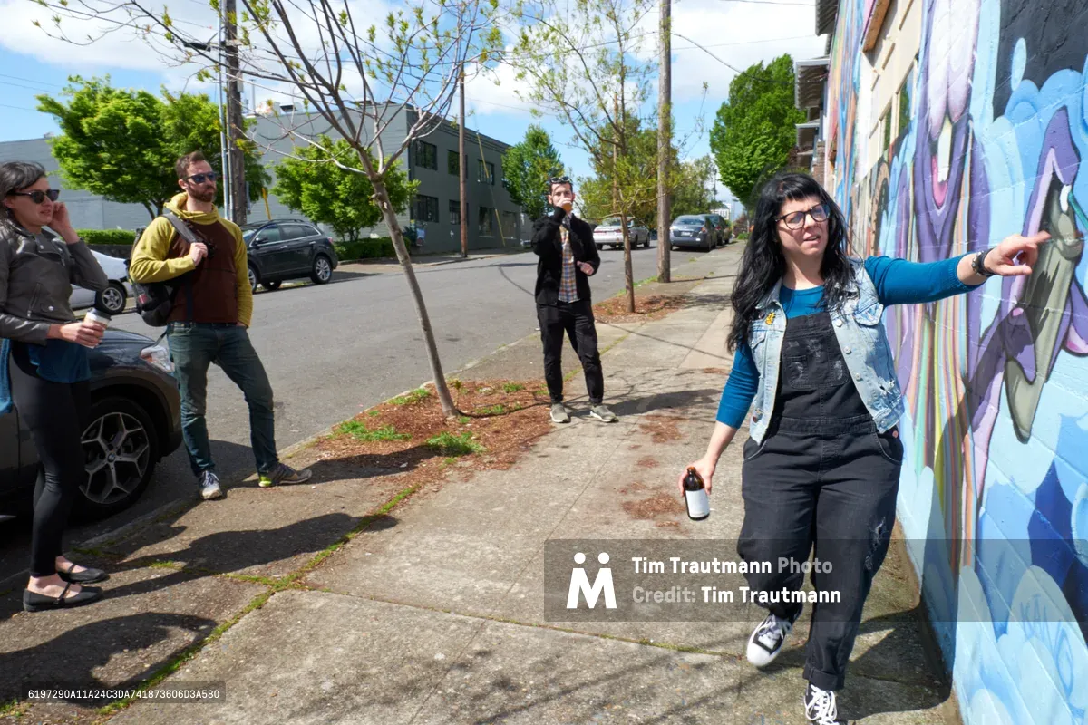Tiffany Conklin of the Portland Street Art Alliance gestures enthusiastically toward a vibrant purple and blue mural while leading an interpretive walking tour in Portland's Central Eastside neighborhood. The afternoon light illuminates the group of engaged participants scattered along the sidewalk, some leaning against cars while others stand attentively. The scene captures the grassroots cultural energy of Southeast Portland's thriving street art community, with newly planted street trees and residential buildings forming the backdrop.