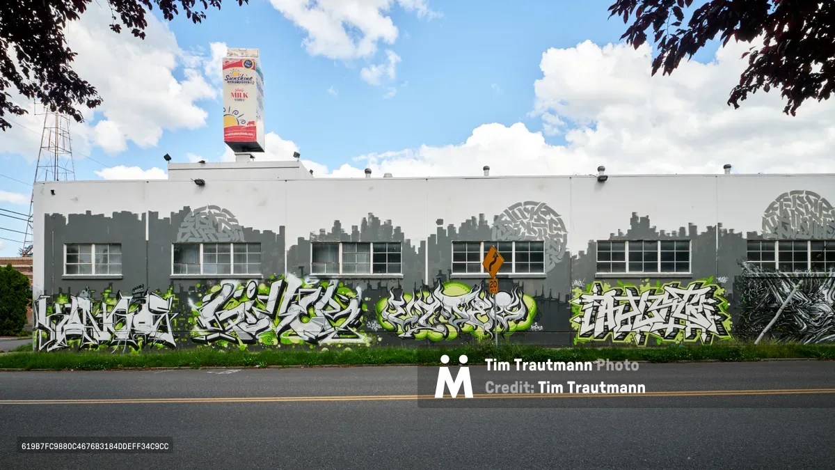A sprawling white industrial building along Northeast 21st Avenue in Portland's Kerns neighborhood displays vibrant green and white graffiti murals across its entire facade. The former Sunshine Dairy site, crowned with a vintage milk carton sign, stands beneath dramatic cloudy skies while street art transforms the utilitarian architecture into an urban gallery. Dark maple foliage frames the scene, creating a natural border around this collision of industrial heritage and contemporary street culture.