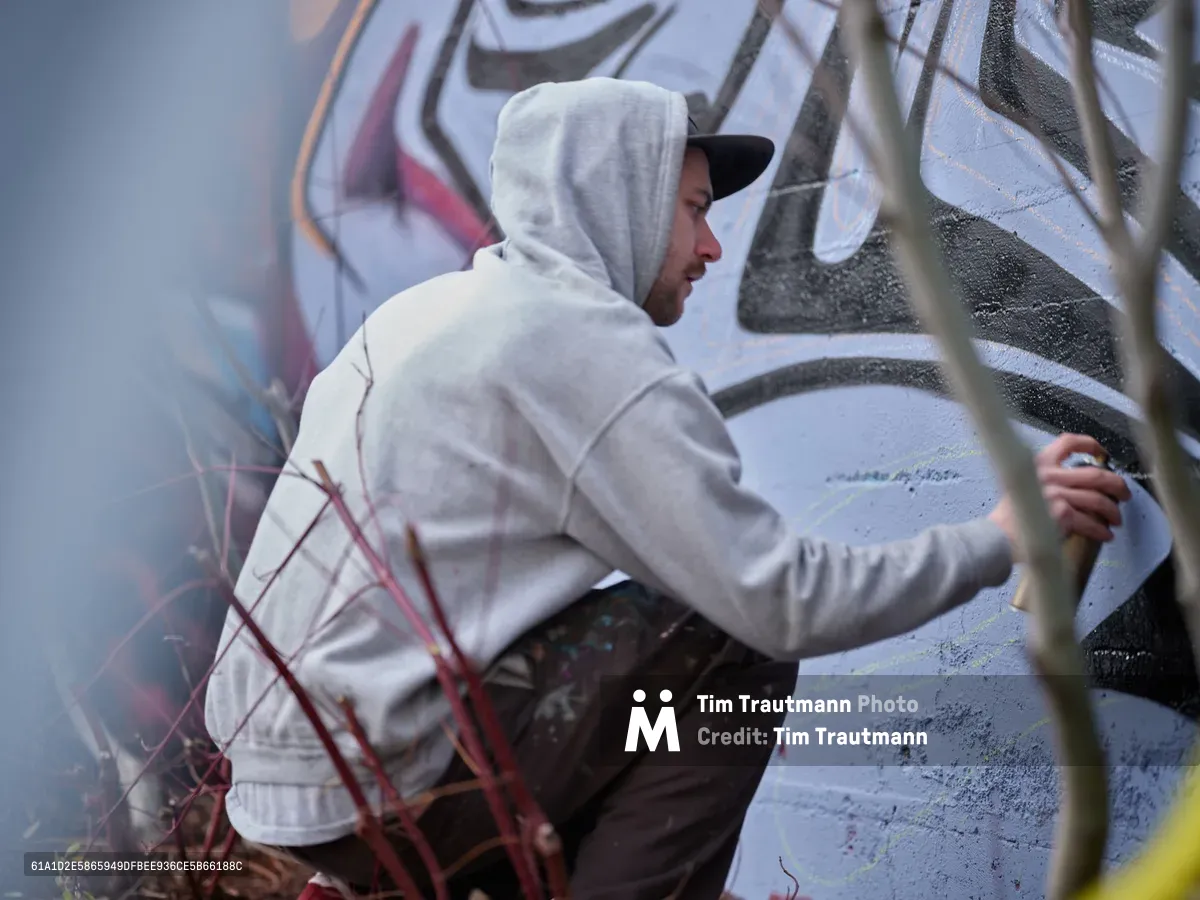 Travis Fields, known as Campographic, works intently on a large-scale mural at the Clay Creative Building in southeast Portland's Hosford-Abernethy neighborhood. Dressed in a light hoodie with hood up and black cap, the artist applies paint to the concrete wall while surrounded by urban textures and overgrown vegetation. The industrial setting and focused concentration capture the authentic spirit of Portland's thriving street art scene.
