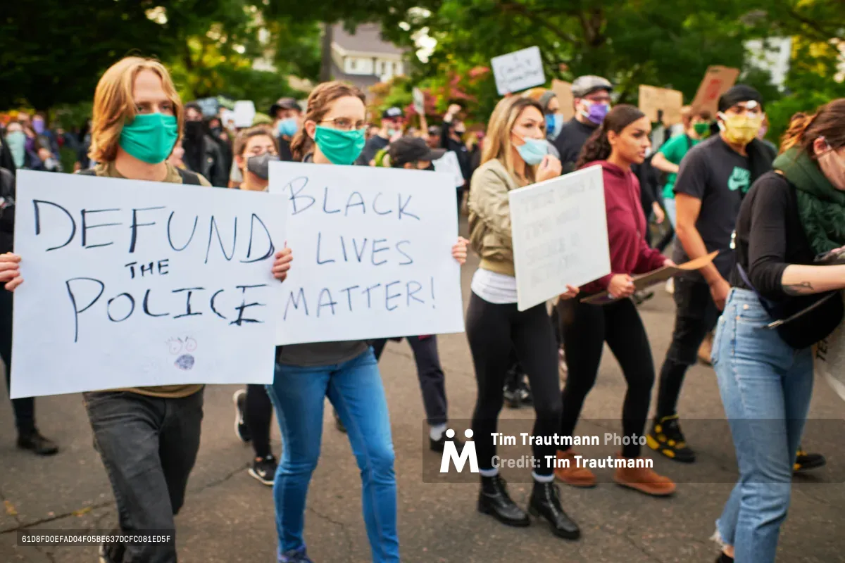 Masked protesters march through tree-lined residential streets of Northeast Portland, their handwritten signs demanding police defunding and affirming that Black lives matter. The diverse crowd moves with quiet determination beneath a canopy of summer foliage, their colorful face coverings and scattered banners creating a tapestry of resistance against the backdrop of suburban homes. The dappled afternoon light filters through leaves, casting the scene in the golden hour glow that defined countless demonstrations during the summer of 2020.