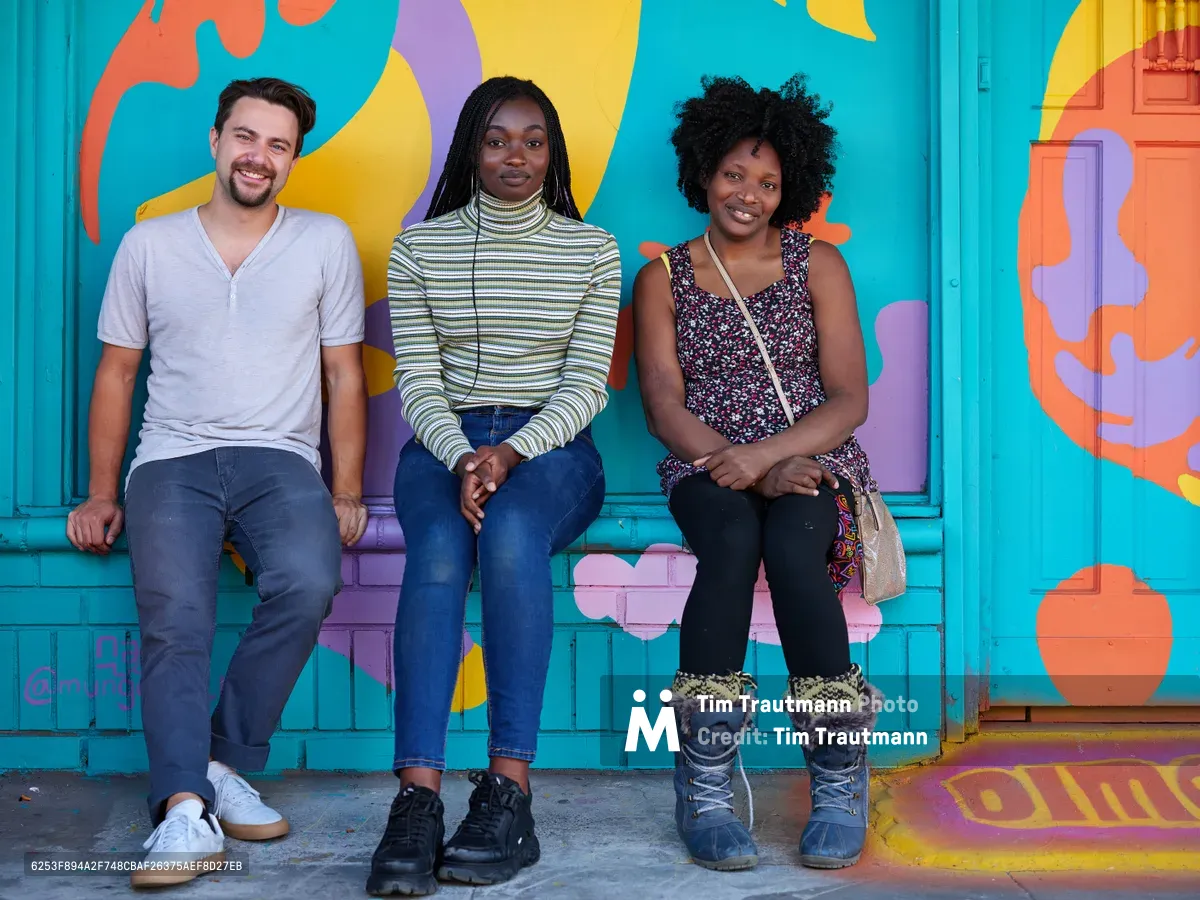 Travis Fields, Naomi Likayi, and the owner of Dime & Penny sit together against a vibrant turquoise wall adorned with flowing abstract murals in coral, yellow, and purple. The trio embodies the creative spirit of Portland's Alberta Arts District, with warm natural light illuminating their relaxed poses and genuine expressions. Their casual yet distinct personal styles—from Fields' henley and jeans to Likayi's striped turtleneck and the business owner's patterned dress—reflect the neighborhood's blend of artistic authenticity and entrepreneurial energy.