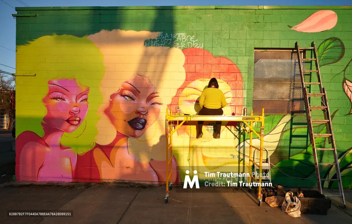 Multidisciplinary artist Villatose works from a yellow scissor lift, adding finishing touches to 'Jardín Cubana,' a stunning large-scale mural featuring three female figures with voluminous natural hair rendered in brilliant yellows, pinks, and corals. The vibrant street art transforms a green concrete block wall on Northeast Alberta Street in Portland's celebrated Alberta Arts District, where golden afternoon light bathes both the artist and their colorful creation. Painting supplies and equipment rest at the base of the wall, capturing the authentic working process of contemporary urban muralism.