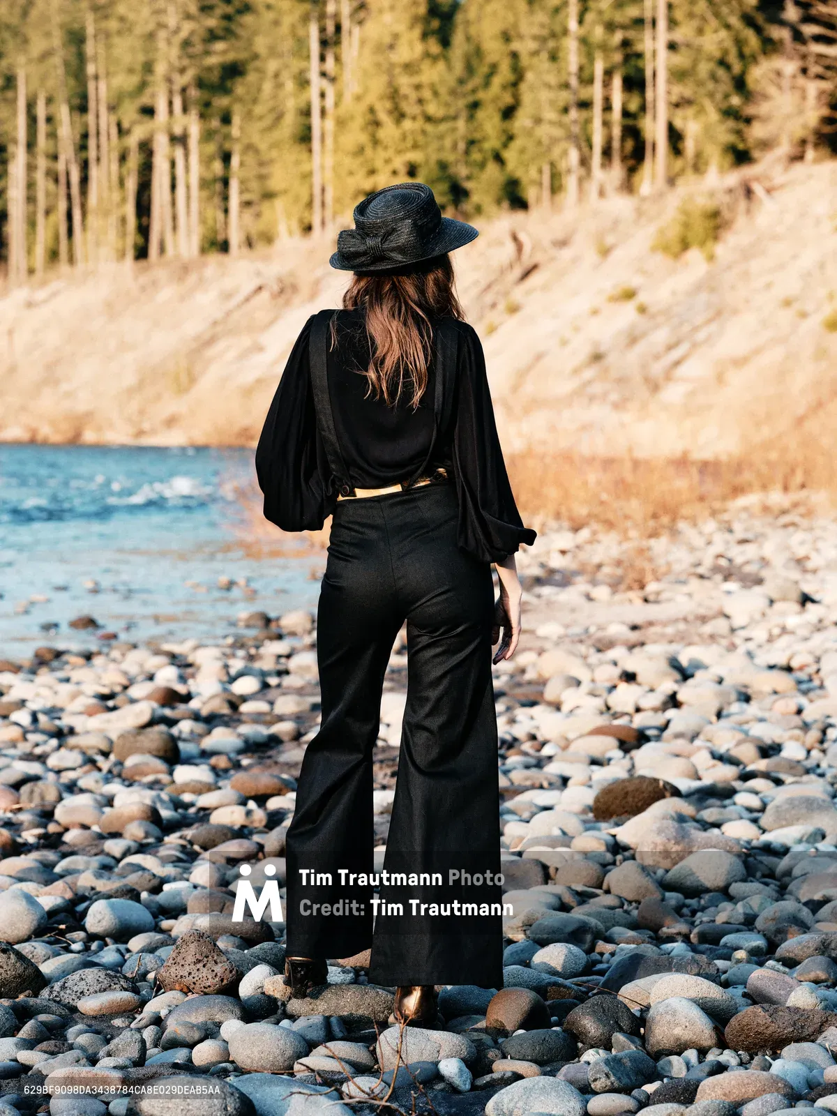 A lone woman in flowing black attire stands with her back to the viewer on the rocky shoreline of the Columbia River along Oregon's Floodplain Trail. Her wide-brimmed hat and billowing sleeves create dramatic silhouettes against the warm afternoon light that bathes the river stones and towering forest beyond. The scene captures a moment of quiet contemplation, with smooth stones of the riverbed stretching toward gentle waters while dense Pacific Northwest woodland rises from the opposite bank.