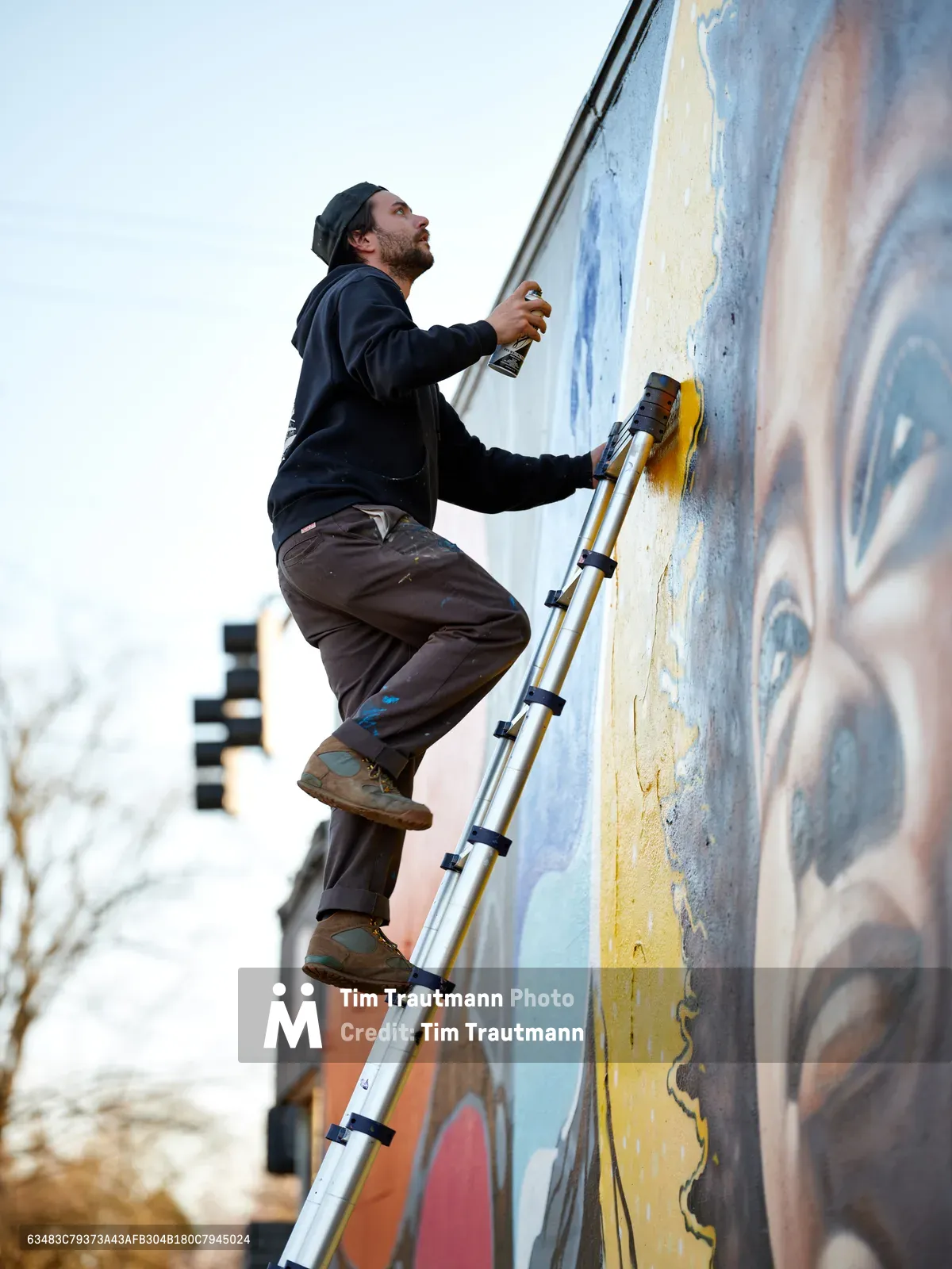 Street artist Travis Fields, working under the moniker campographic, balances precariously on an aluminum ladder as he spray paints details of poet Amanda Gorman's portrait on a building wall at Northeast 15th and Killingsworth in Portland's Alberta district. The bearded artist in dark clothing focuses intently on his work against the backdrop of pastel blues, warm oranges, and golden yellows that form the emerging mural. Urban buildings blur softly in the background under overcast Pacific Northwest skies, capturing a moment of artistic creation transforming the neighborhood streetscape.