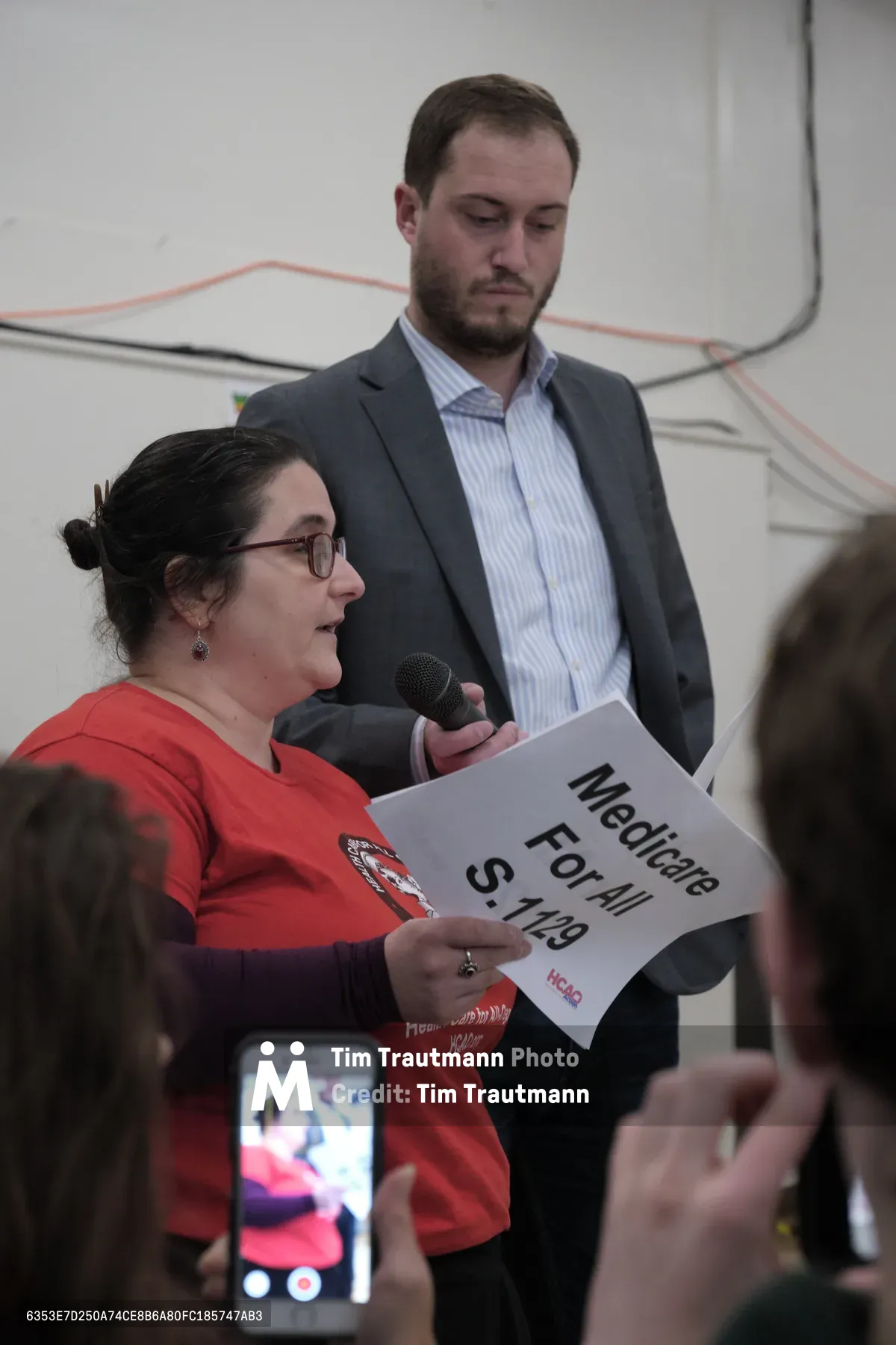 At Robert Gray Middle School in Portland's Hillsdale neighborhood, a woman in a vibrant red shirt holds a "Medicare For All S.149" sign while speaking into a microphone during Senator Ron Wyden's town hall meeting. A suited man stands attentively behind her as smartphone screens capture the moment from the audience below. The institutional setting with its neutral walls and orange cable conduits frames this moment of grassroots political engagement.
