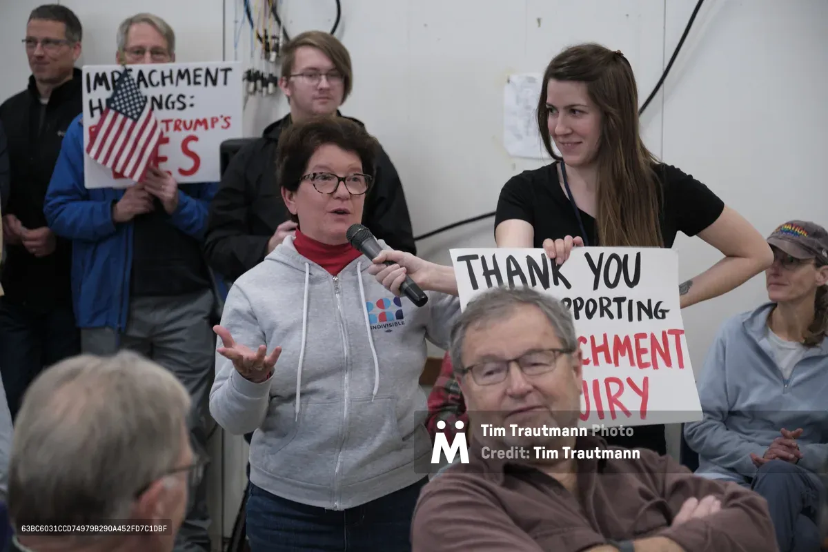 A woman in a gray hoodie addresses Senator Ron Wyden through a microphone at a crowded town hall meeting in Robert Gray Middle School's gymnasium. Behind her, constituents hold handmade signs reading "IMPEACHMENT HEARINGS: TRUMP'S LIES" and "THANK YOU FOR SUPPORTING IMPEACHMENT INQUIRY," their faces reflecting the charged political atmosphere. The stark institutional lighting and white-walled setting emphasize the grassroots nature of this democratic engagement in Portland's Hillsdale neighborhood.