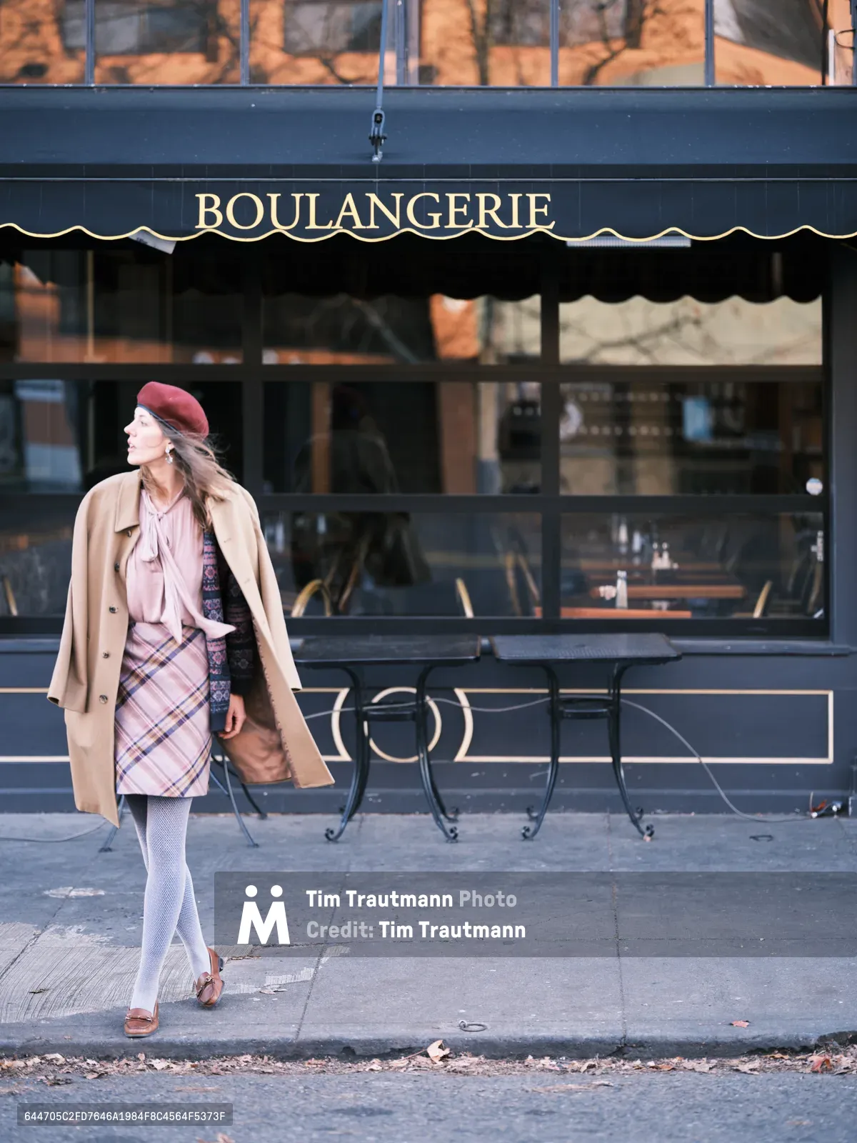 A woman in a burgundy beret and camel trench coat strolls past a classic French boulangerie on Northeast Alberta Street in Portland, Oregon. Her layered outfit features a pink plaid skirt, rose-toned blouse, and white tights, creating an effortlessly chic ensemble. The golden hour light bathes the blue storefront and sidewalk in warm amber tones, while autumn leaves scatter across the pavement, capturing the romantic intersection of French café culture and Pacific Northwest charm.
