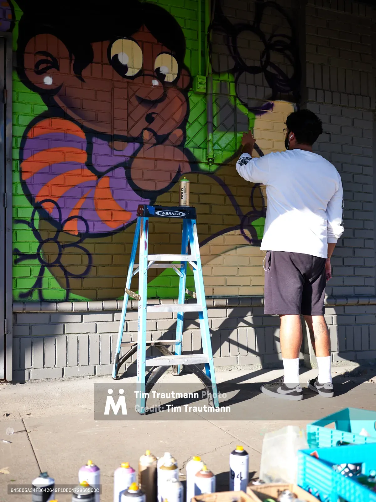 Muralist Heysus works methodically on a colorful character-based mural along the brick wall of a building in Portland's Alberta neighborhood. The artist, dressed casually in a white long-sleeve shirt and gray shorts, stands on a blue Werner ladder surrounded by an array of spray paint cans scattered across the concrete sidewalk. His emerging artwork features a whimsical monkey character rendered in warm earth tones against brilliant lime green backgrounds, capturing the dynamic spirit of Northeast Portland's thriving street art scene.