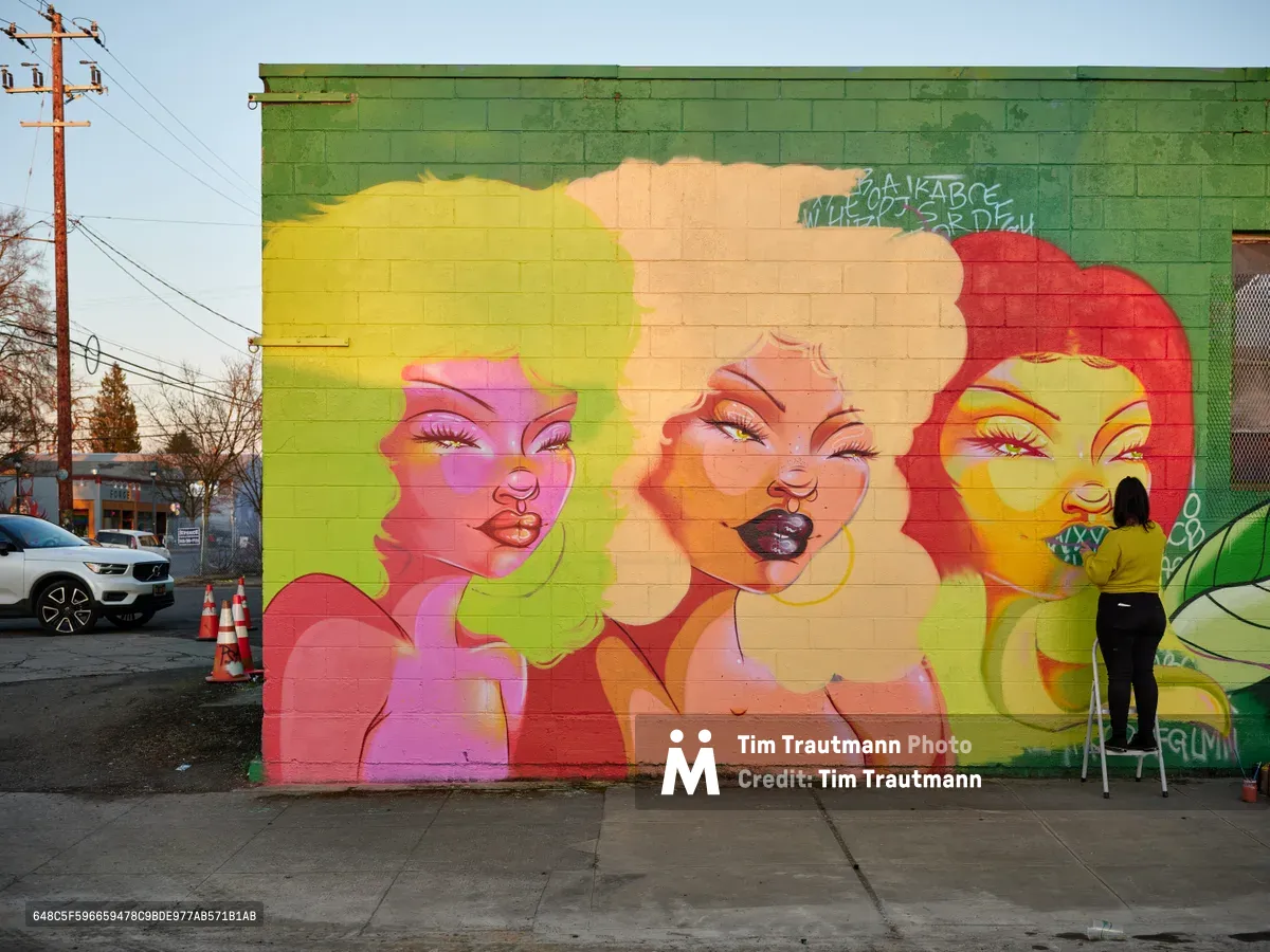 Multidisciplinary artist Villatose applies finishing touches to Jardín Cubana, a striking mural featuring three colorful female portraits on a green cinderblock wall in Portland's Alberta Arts District. The artist, positioned on a small stepladder, works with concentrated focus while the late afternoon light illuminates the vivid yellow, pink, and coral faces that emerge from the urban canvas. Traffic cones and power lines frame this moment of artistic creation in the heart of Northeast Portland's creative corridor.
