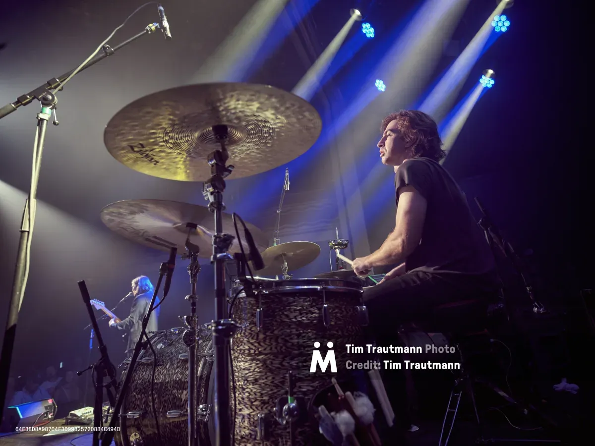 Julian Dorio commands the stage behind his drum kit during Band of Skulls' 10 Year Anniversary Tour at Webster Hall in Manhattan. Theatrical blue stage lights pierce through atmospheric haze, creating dramatic shafts of illumination that frame the focused drummer in profile. The intimate venue setting captures the raw energy of live performance, with gleaming cymbals suspended overhead and a bassist visible in the background, all enveloped in the moody ambiance of concert lighting.