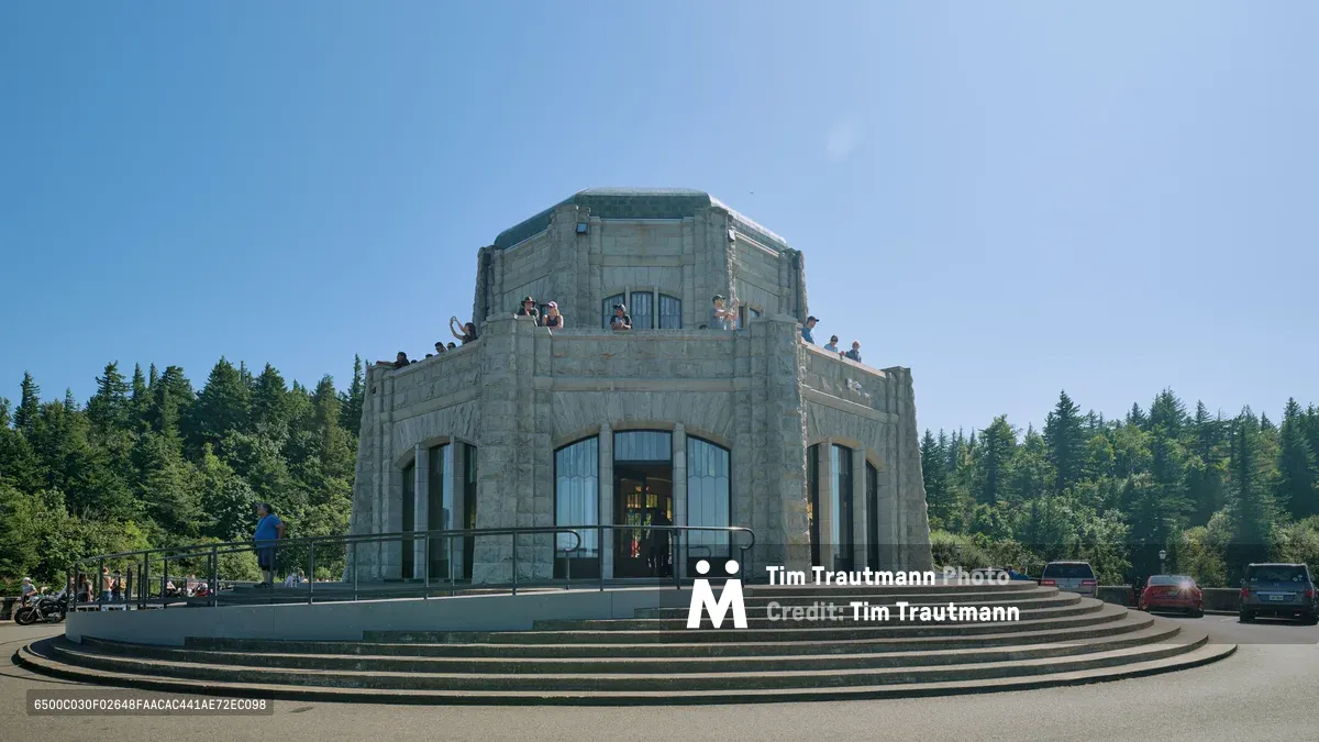 The iconic Vista House stands majestically against a brilliant azure sky at Crown Point, its Art Deco limestone facade gleaming in golden afternoon light. Visitors gather on the observation deck's upper levels, silhouetted against the vast expanse above the Columbia River Gorge. The octagonal structure's neoclassical columns and curved stonework create dramatic shadows across the sweeping circular steps, while evergreen forests frame this architectural monument to Oregon's scenic highway heritage.